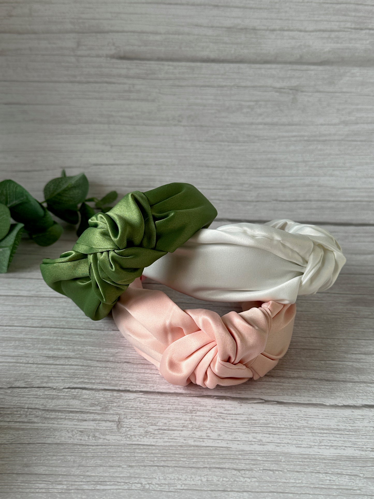 Displayed against a light wooden background are three handcrafted Satin Knotted Headbands from Crafts by Kate's Bridal Collection. The headbands, available in green, white, and pink, are accented with a few green leaves to bring a natural touch to the scene.
