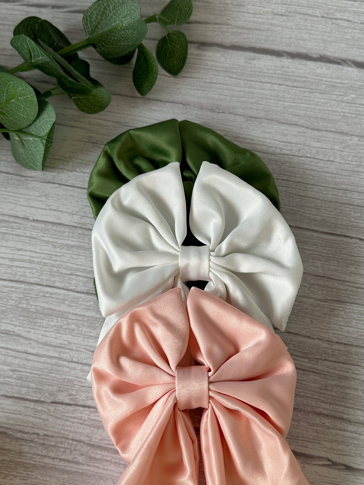 Three satin scrunchies from the Crafts by Kate Bridal Collection, each adorned with large sailor bows, are stacked on a light wooden surface. From top to bottom, the Satin Sailor Bow with hair band is available in green, white, and peach. A green plant with round leaves is partially visible in the upper left corner, adding a touch of handcrafted luxury to the scene.