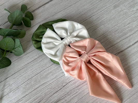 A large green Satin Sailor Bow with a hair band from our Handcrafted Luxury Bridal Collection by Crafts by Kate rests on a light wood surface. To the upper left of the bow, a sprig of eucalyptus leaves adds a touch of nature to the scene. The bow's silky fabric catches the light, highlighting its elegant, smooth texture—a perfect piece for any bride-to-be.
