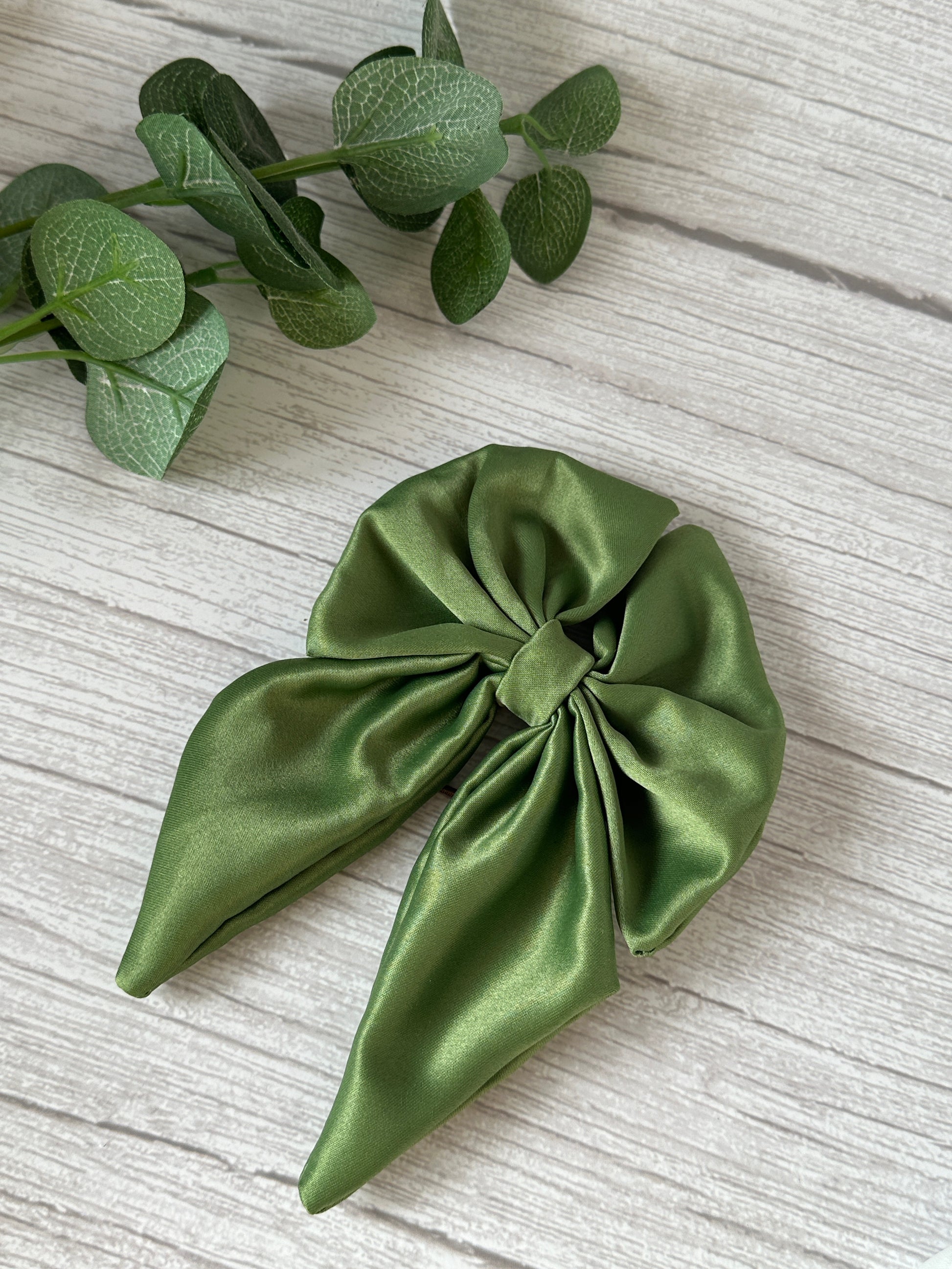 A large green Satin Sailor Bow with a hair band from our Handcrafted Luxury Bridal Collection by Crafts by Kate rests on a light wood surface. To the upper left of the bow, a sprig of eucalyptus leaves adds a touch of nature to the scene. The bow's silky fabric catches the light, highlighting its elegant, smooth texture—a perfect piece for any bride-to-be.