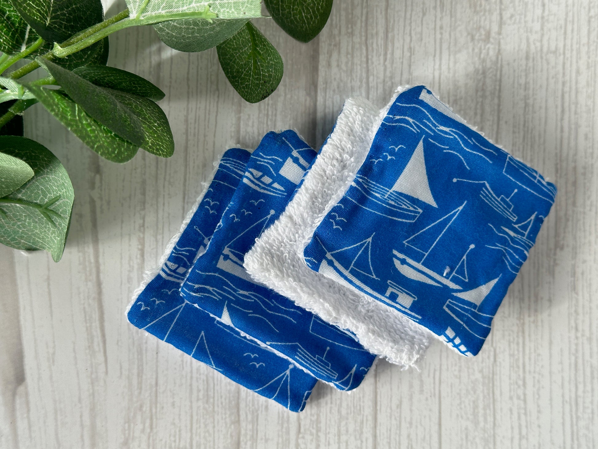 Four Cotton Reusable Make-up Remover Pads - Seaside Blue by Crafts by Kate, stack on a light wooden surface with white nautical patterns of sailboats and waves. They are crafted from sustainable materials, with a green leafy plant partially visible on the left side.