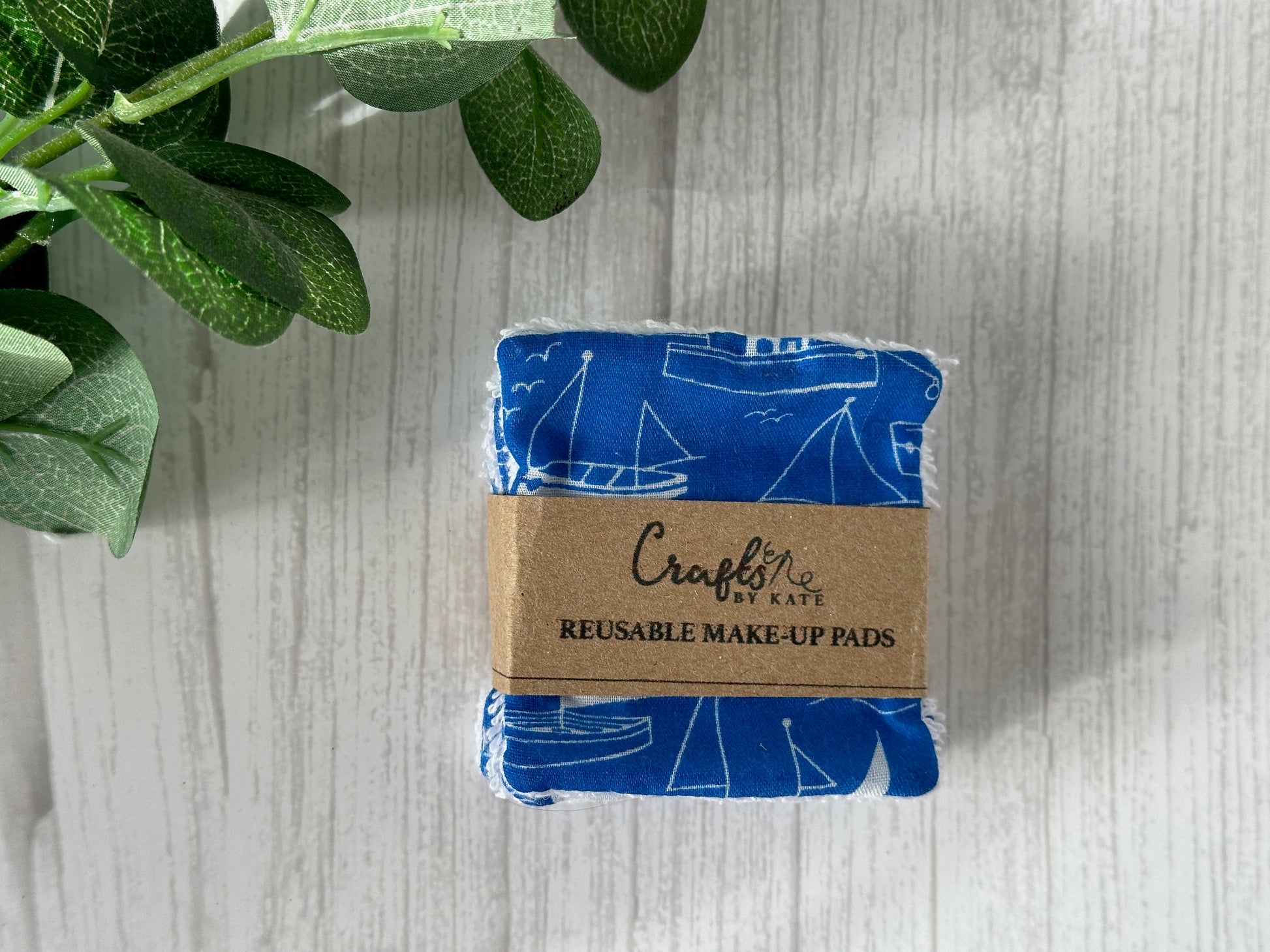 The Cotton Reusable Make-up Remover Pads - Seaside Blue by Crafts by Kate feature a nautical sailboat design. They're stacked and wrapped with a brown paper band labeled "Crafts by Kate," displayed on a light wooden surface next to green leaves, emphasizing their eco-friendly nature.