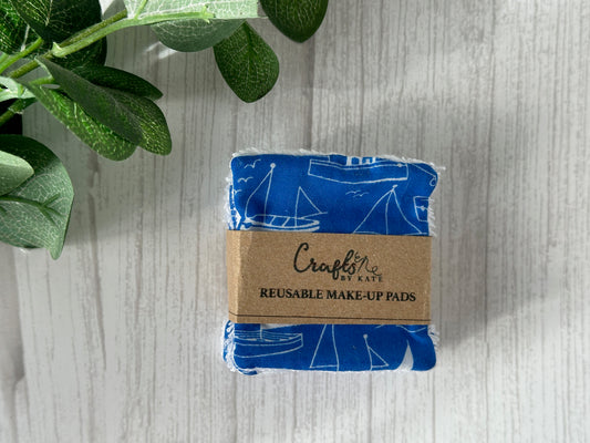 The Cotton Reusable Make-up Remover Pads - Seaside Blue by Crafts by Kate feature a nautical sailboat design. They're stacked and wrapped with a brown paper band labeled "Crafts by Kate," displayed on a light wooden surface next to green leaves, emphasizing their eco-friendly nature.
