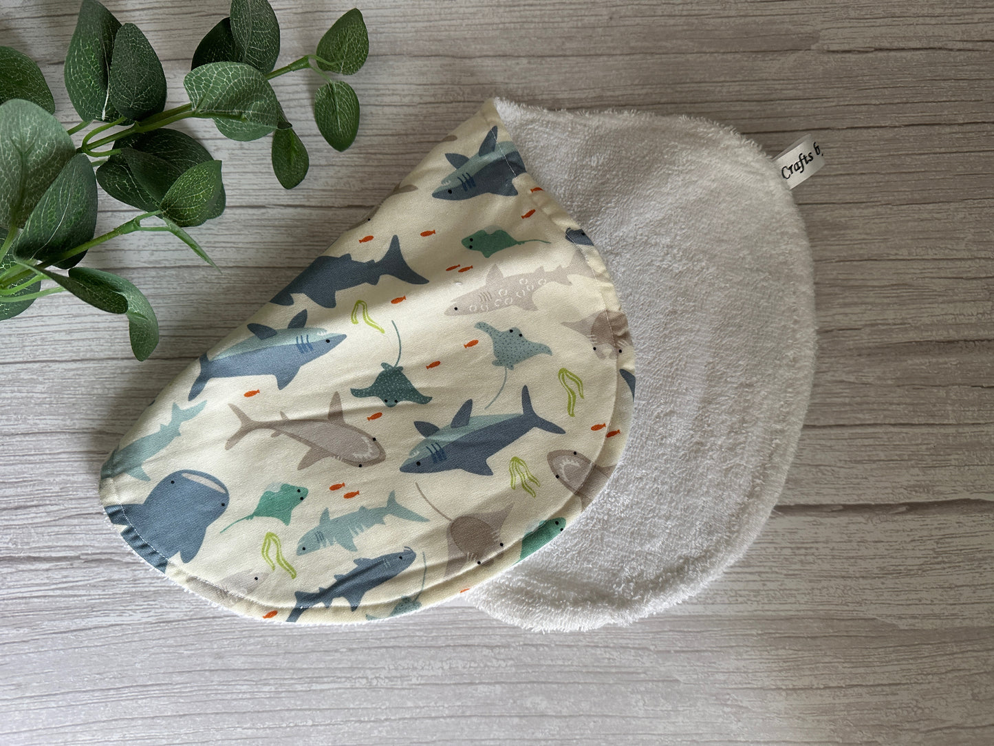 On a wooden surface lies the Cotton & Terry Towelling Baby Burping Cloth - Shark Tank, a semi-circular piece handcrafted by Crafts by Kate. It showcases a shark pattern with various species in muted colors and is made from ultra-soft cotton, ideal for use as a burping cloth for babies. The reverse side is white and textured. A green plant sits beside it.
