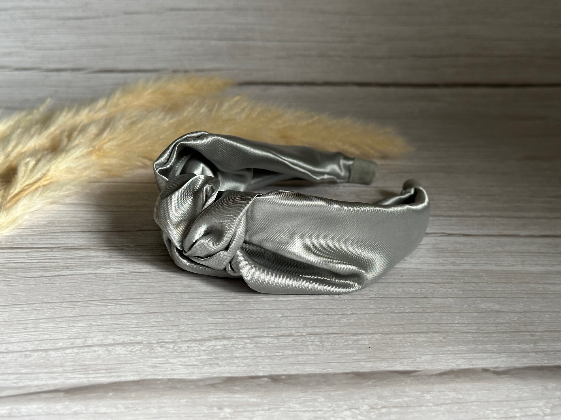 A luxurious Silk Knotted Headband - Metallic Silver from Crafts by Kate sits on a light wooden surface, radiating elegance. In the background, beige pampas grass is partially visible, adding to the scene's sophisticated charm.
