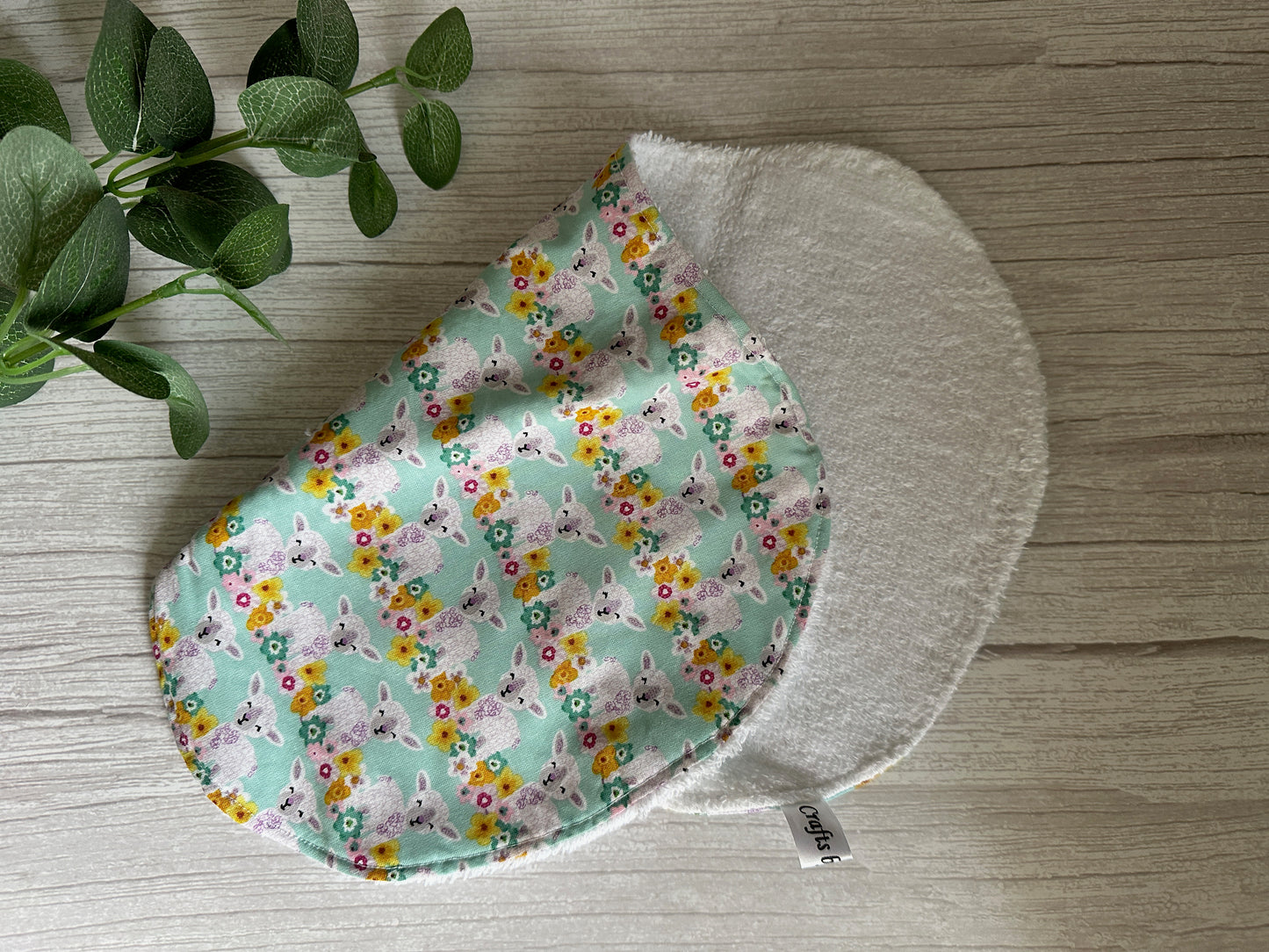 A handcrafted Cotton & Terry Towelling Baby Burping Cloth from Crafts by Kate features a printed design of playful grey cats and colorful flowers on a green background. It rests on a wooden surface next to a small green plant, infusing the scene with the lively charm of spring.