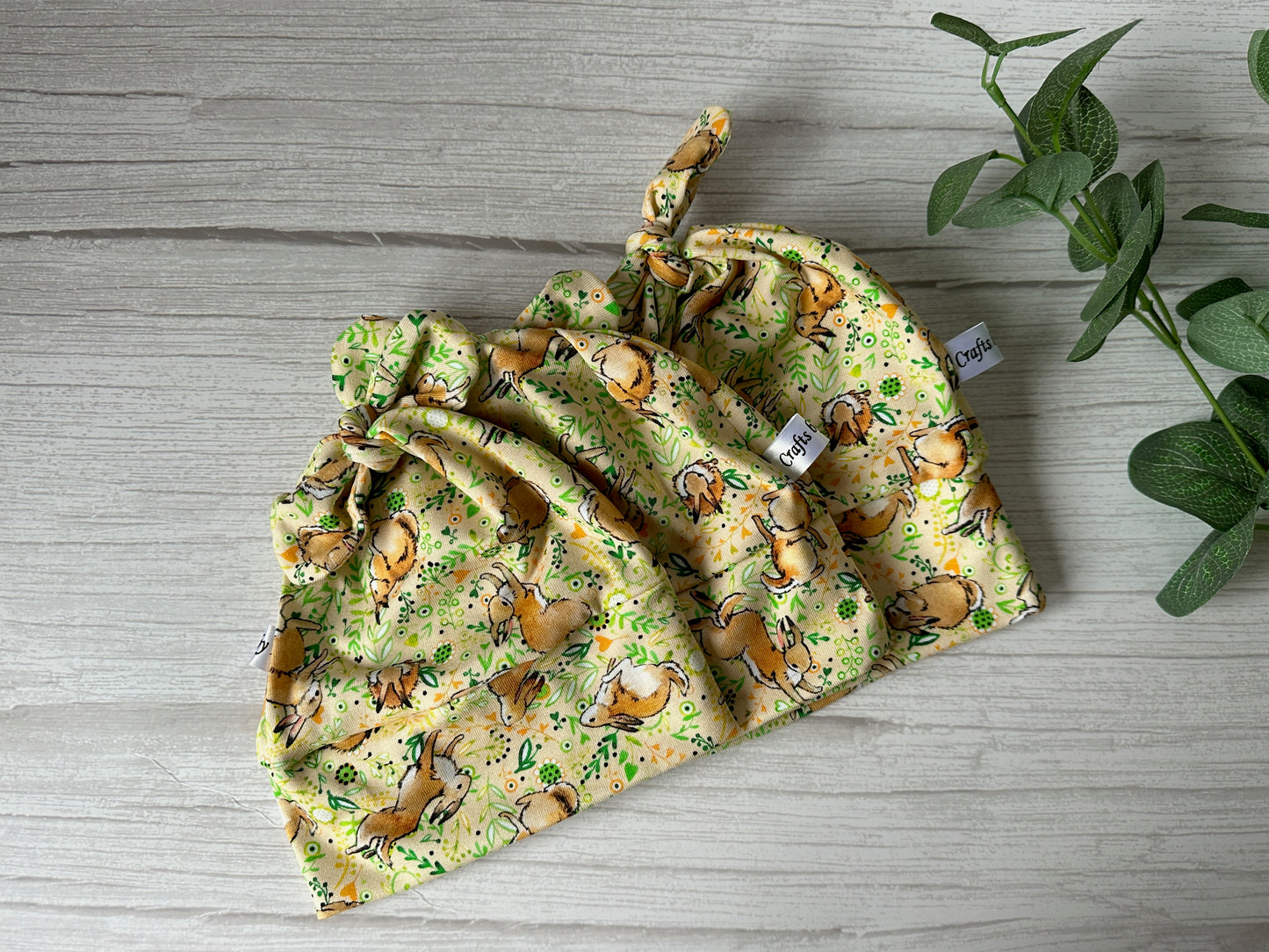 Three yellow "Baby Wool Cotton Jersey Knotted Hats - Spring Rabbits" by Crafts by Kate, featuring whimsical animal and leaf patterns, are displayed on a light wooden surface next to green foliage.