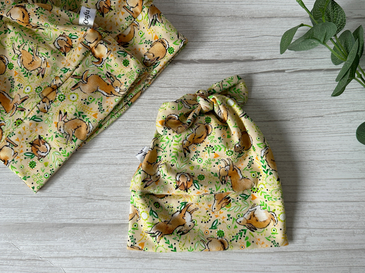 A knotted baby hat from Crafts by Kate and a matching cloth rest on a wooden surface, both made from luxury wool jersey with charming yellow and green spring rabbit and flower patterns. A sprig of green leaves can be seen in the top right corner, adding natural elegance.