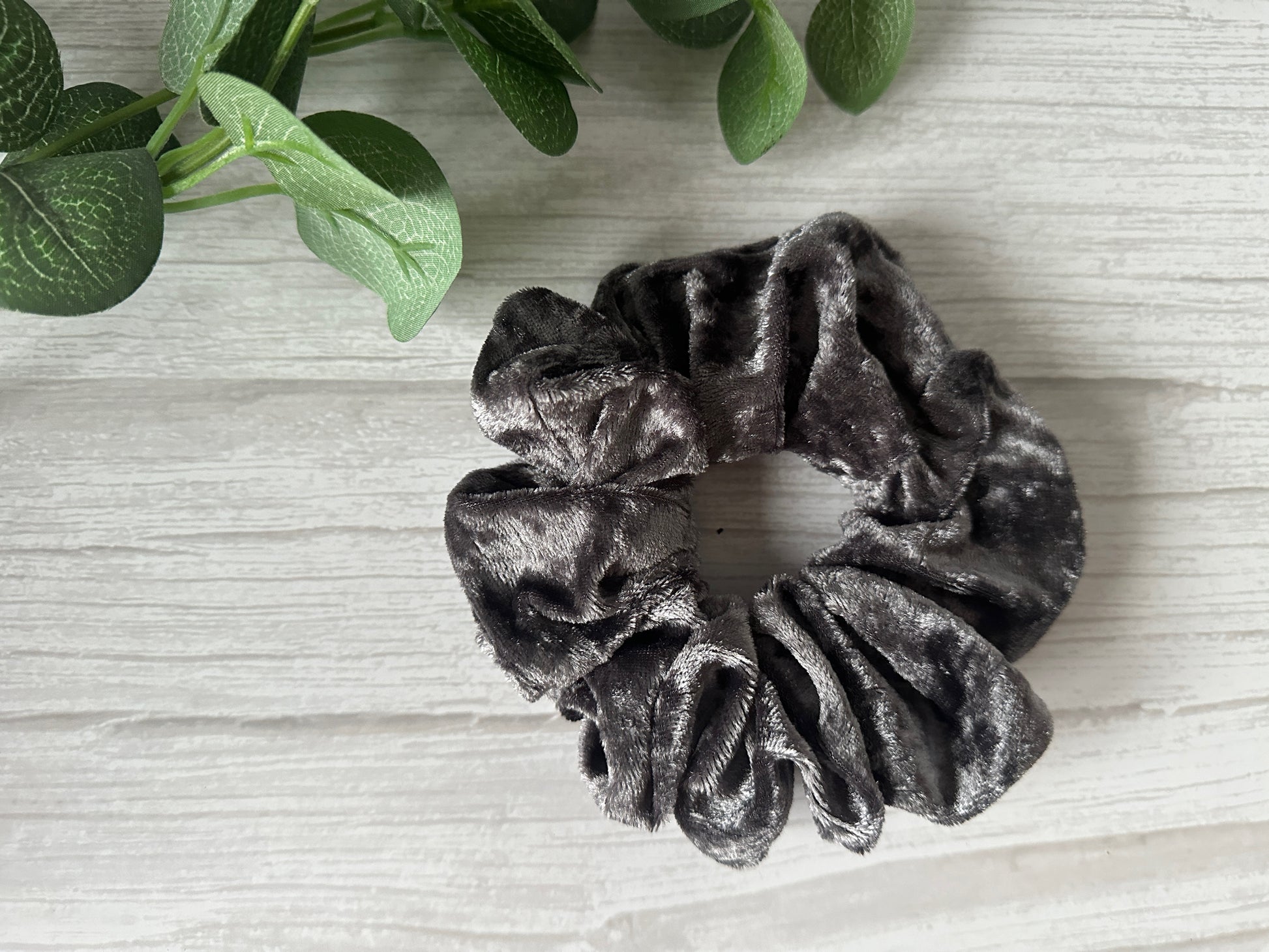 The Crushed Velvet Extra Large Scrunchie in Steel Grey by Crafts by Kate is displayed on a light wooden surface, accented with green leafy plants for a natural touch.