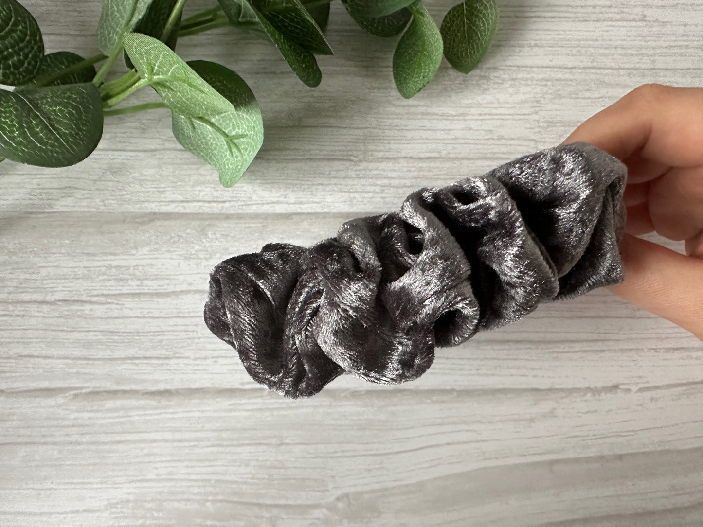 A hand holds the Crafts by Kate Crushed Velvet Extra Large Scrunchie in Steel Grey on a light wooden surface, with green plant leaves visible in the top left corner.
