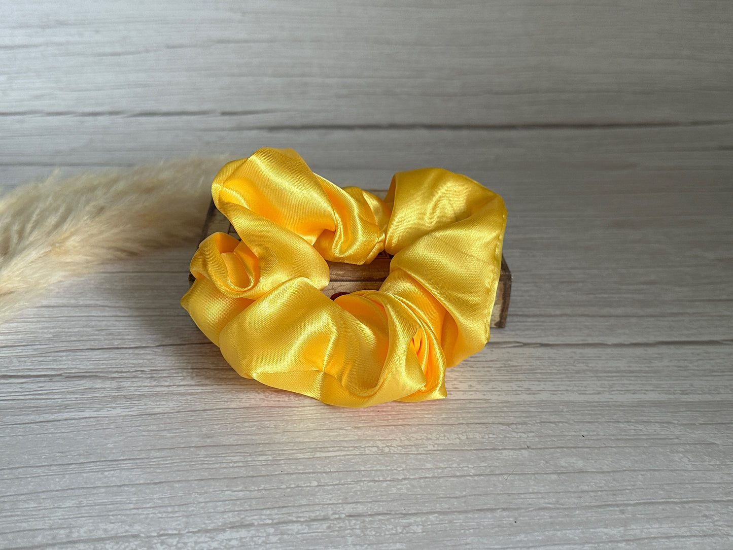 The Silk Extra Large Scrunchie in Sunshine Yellow from Crafts by Kate, featuring a silky texture, is set on a light wooden surface. A fluffy beige pampas grass, blurred in the background, adds a natural touch to the scene.