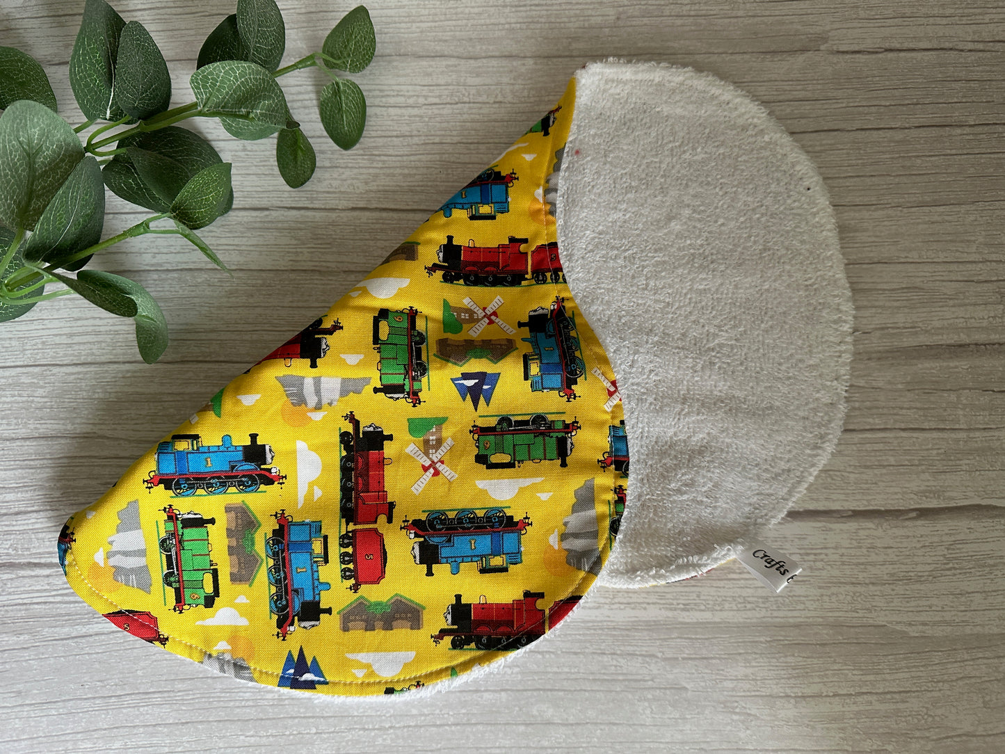 The Cotton & Terry Towelling Baby Burping Cloth - Thomas The Tank Engine by Crafts by Kate, crafted from ultra-soft cotton, boasts a delightful train pattern set against a yellow backdrop. It is neatly folded on a wooden surface, with a small green plant partially visible on the left side, enhancing the natural charm of this handcrafted item.