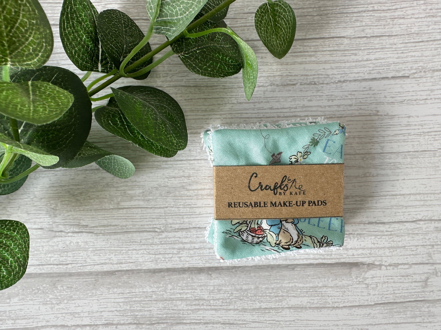 Crafts by Kate Cotton Reusable Make-up Remover Pads in turquoise with Peter Rabbit prints are set on a textured light gray surface, wrapped in a brown label. A leafy green plant is partially visible on the left, highlighting these eco-friendly pads.