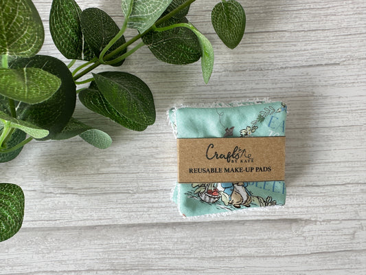 Crafts by Kate Cotton Reusable Make-up Remover Pads in turquoise with Peter Rabbit prints are set on a textured light gray surface, wrapped in a brown label. A leafy green plant is partially visible on the left, highlighting these eco-friendly pads.
