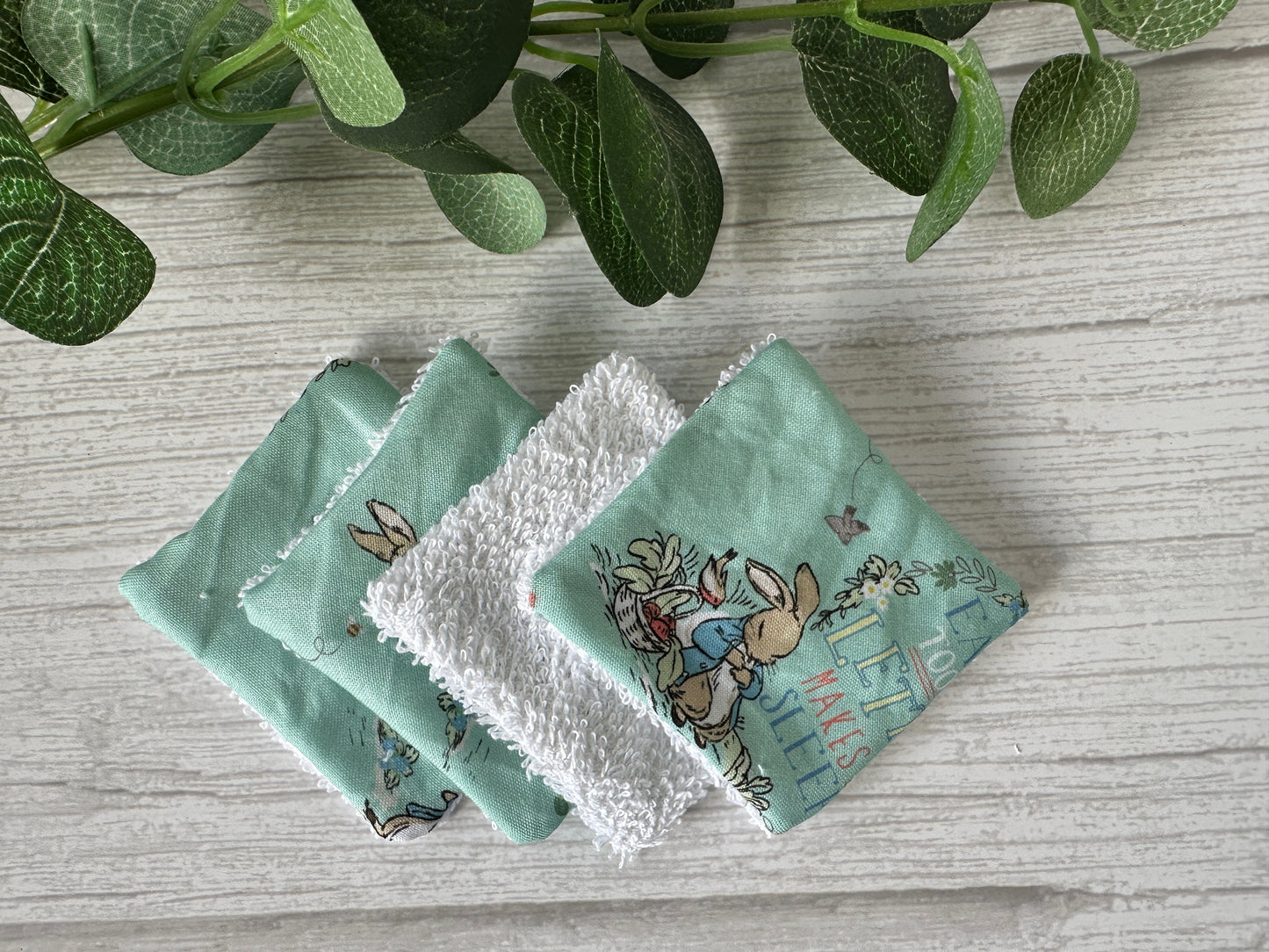 Crafts by Kate's Cotton Reusable Make-up Remover Pads - Peter Rabbit (turquoise) are perfect eco-friendly solutions. These five pastel green and white fabric squares, some with rabbit illustrations, add a sustainable touch to your routine when placed on a light wooden surface with green leaves nearby.