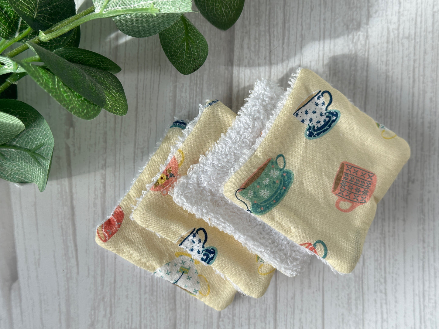 Crafts by Kate's Cotton Reusable Make-up Remover Pads in Yellow Tearoom design lie on a white wooden surface for an eco-friendly look. A small green plant adds nature in the top left corner, with gentle shadows from soft lighting.
