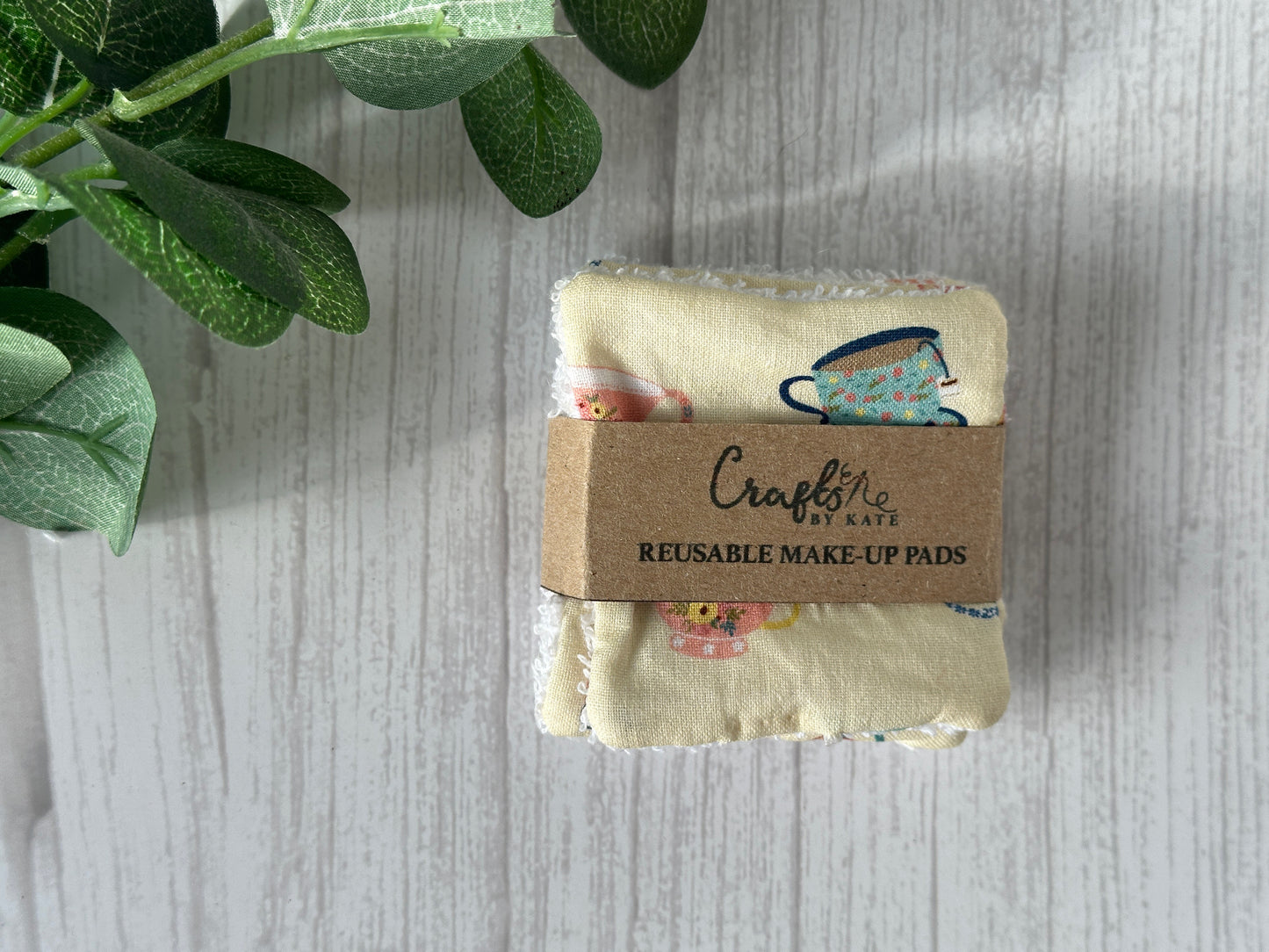 A set of Crafts by Kate Cotton Reusable Make-up Remover Pads - Yellow Tearoom, wrapped in a brown label, rests on a light wooden surface. The sustainable pads display colorful cups and flowers, while a green leafy plant in the top left corner highlights their eco-friendly appeal.