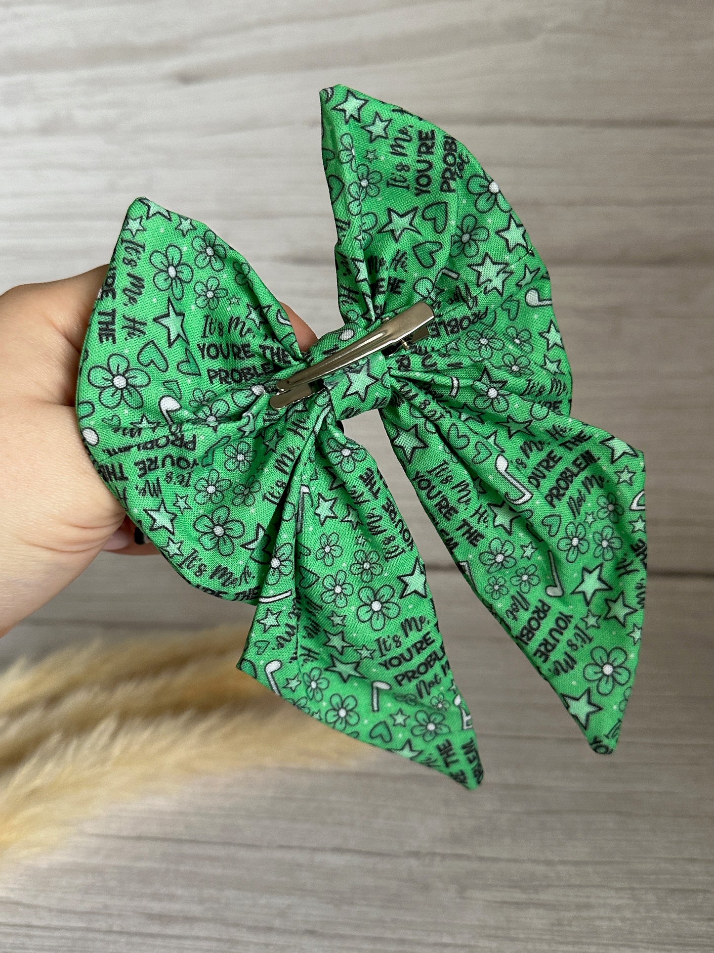 A hand holds the Cotton Sailor Bow Clip - You're the Problem by Crafts by Kate, a large green accessory with floral and star patterns and the phrase "It’s me, hi, I’m the problem, it’s me," shown against a light wood background.