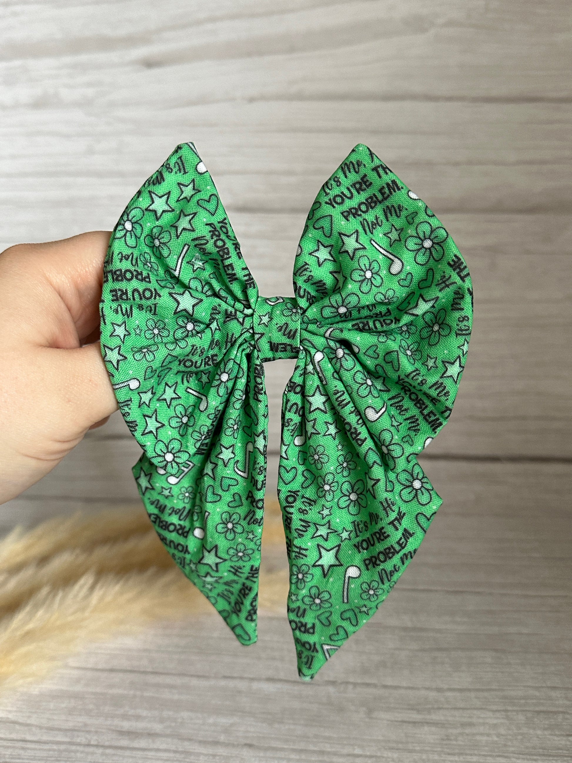 A hand holds the Crafts by Kate Cotton Sailor Bow Clip – "You're the Problem," featuring green cotton with black doodles of stars, flowers, and text on a light wooden background, showcasing handcrafted luxury and a secure crocodile clip.