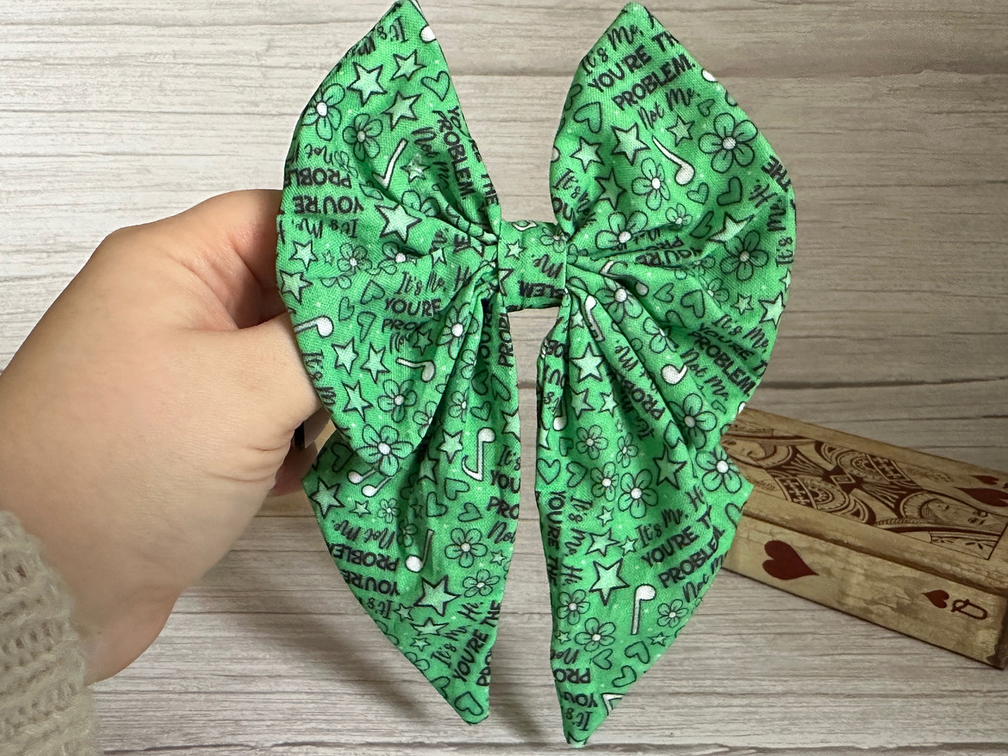 A hand holds the "You're the Problem" Cotton Sailor Bow Clip by Crafts by Kate, a handcrafted luxury cotton accessory with black doodles of stars, flowers, and music notes. A wooden box with heart designs is partially visible in the background.