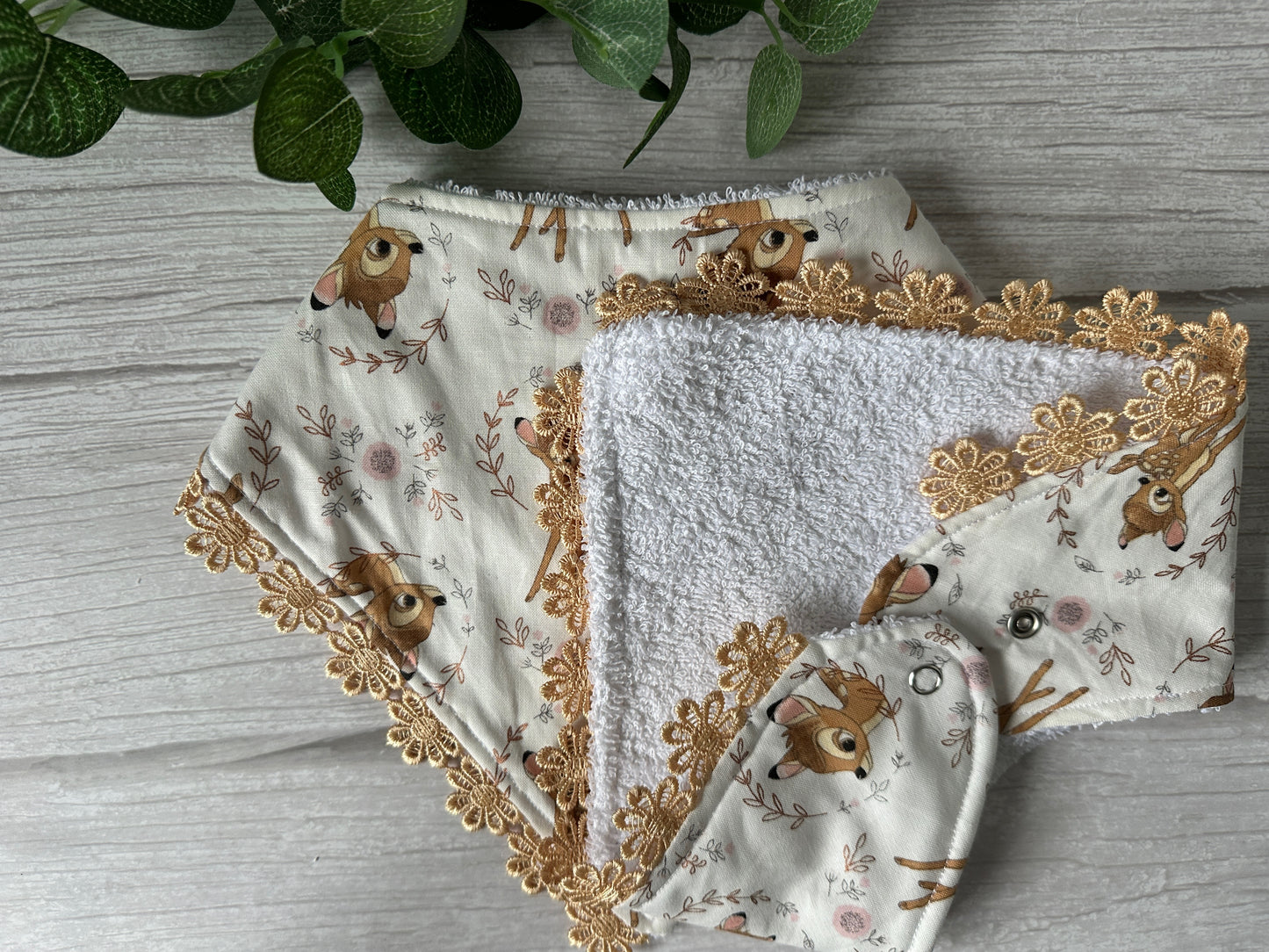 Two cloth diapers with a deer and floral pattern on a light background, brown lace trim, and one open to show its soft white lining. A green leafy plant is partially visible beside Crafts by Kate's handcrafted Cotton & Terry Towelling Baby Bandana Bib - Disney's Bambi with Lace Ribbon Embroidery.