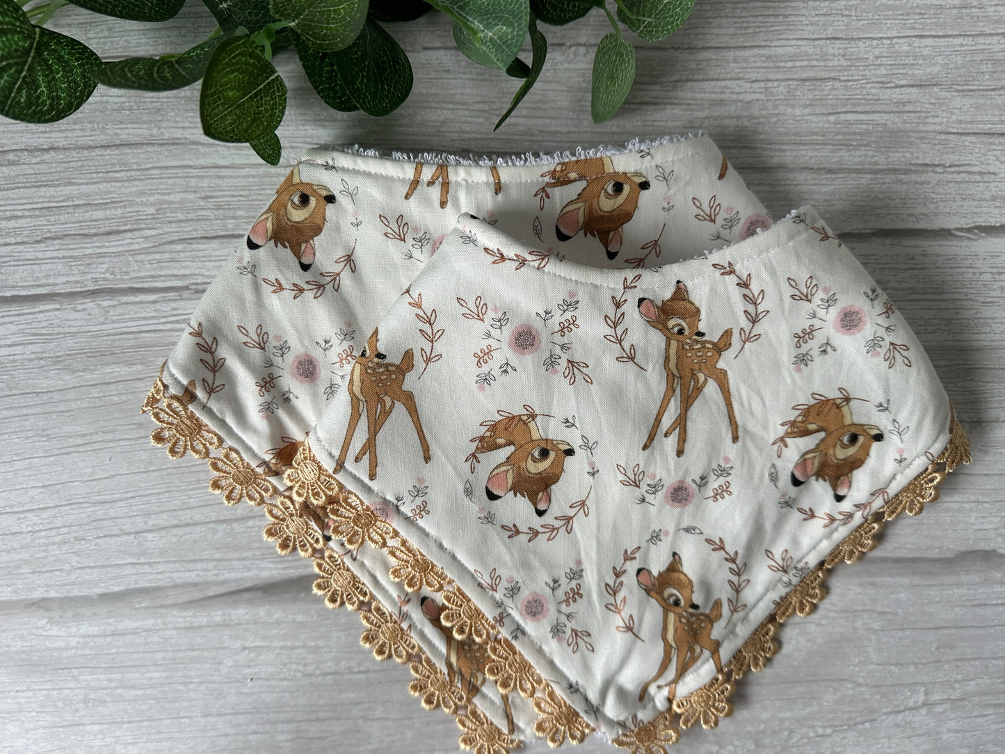 Two Crafts by Kate "Cotton & Terry Towelling Baby Bandana Bibs" featuring Disney's Bambi and lace ribbon embroidery are elegantly placed on a light wood surface, beneath green leaves.