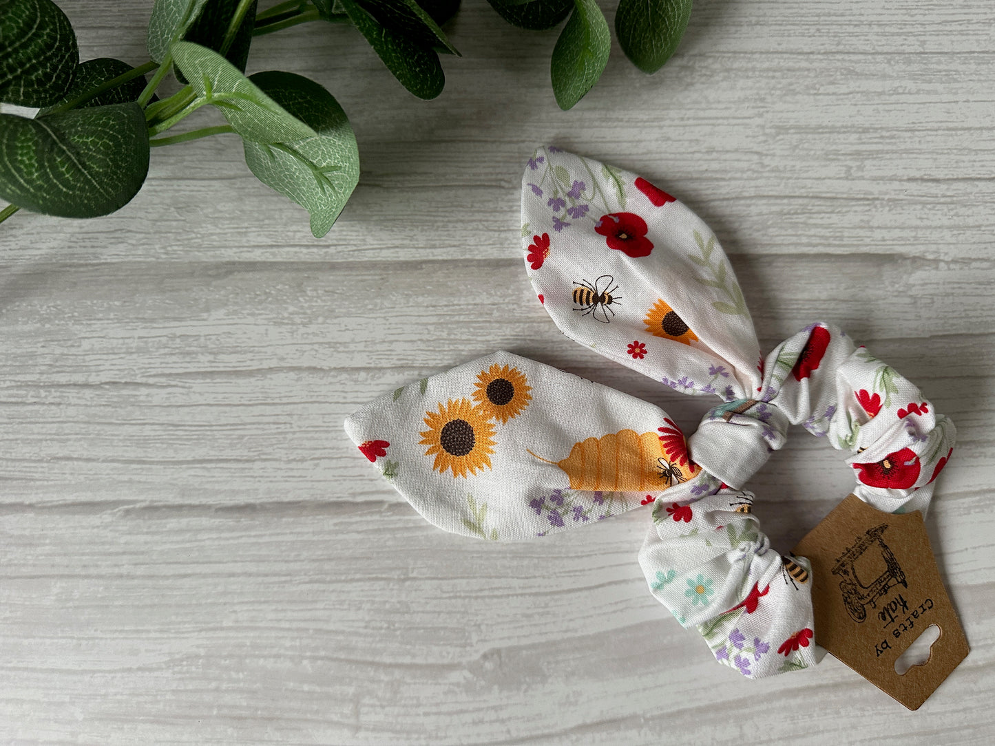 A "Crafts by Kate" luxury "Cotton Scrunchie with Tails - Bumble Bee Garden" sits on a light wooden surface, surrounded by green leaves. Its elegant appeal is enhanced by a small brown tag with text.