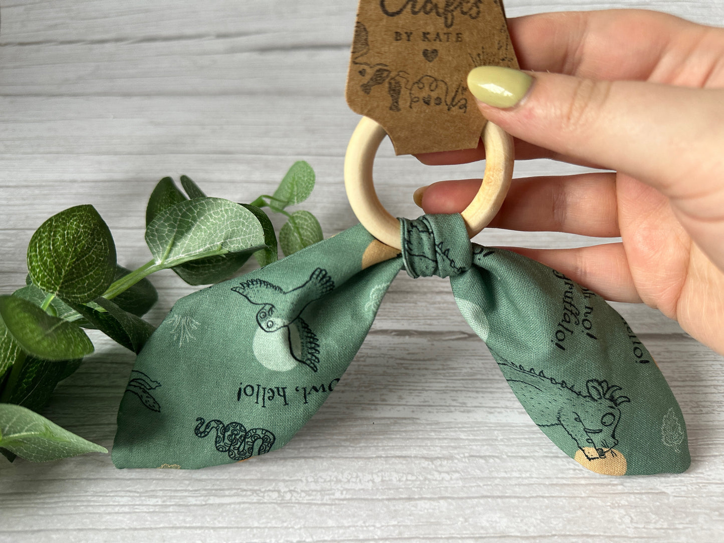 A hand with light green nail polish holds the Cotton & Bamboo Baby Teething Rattle - Green Gruffalo by Crafts by Kate, featuring teal fabric with snake, bird, and rabbit illustrations. To the left, vibrant green leaves rest on a light wooden surface.
