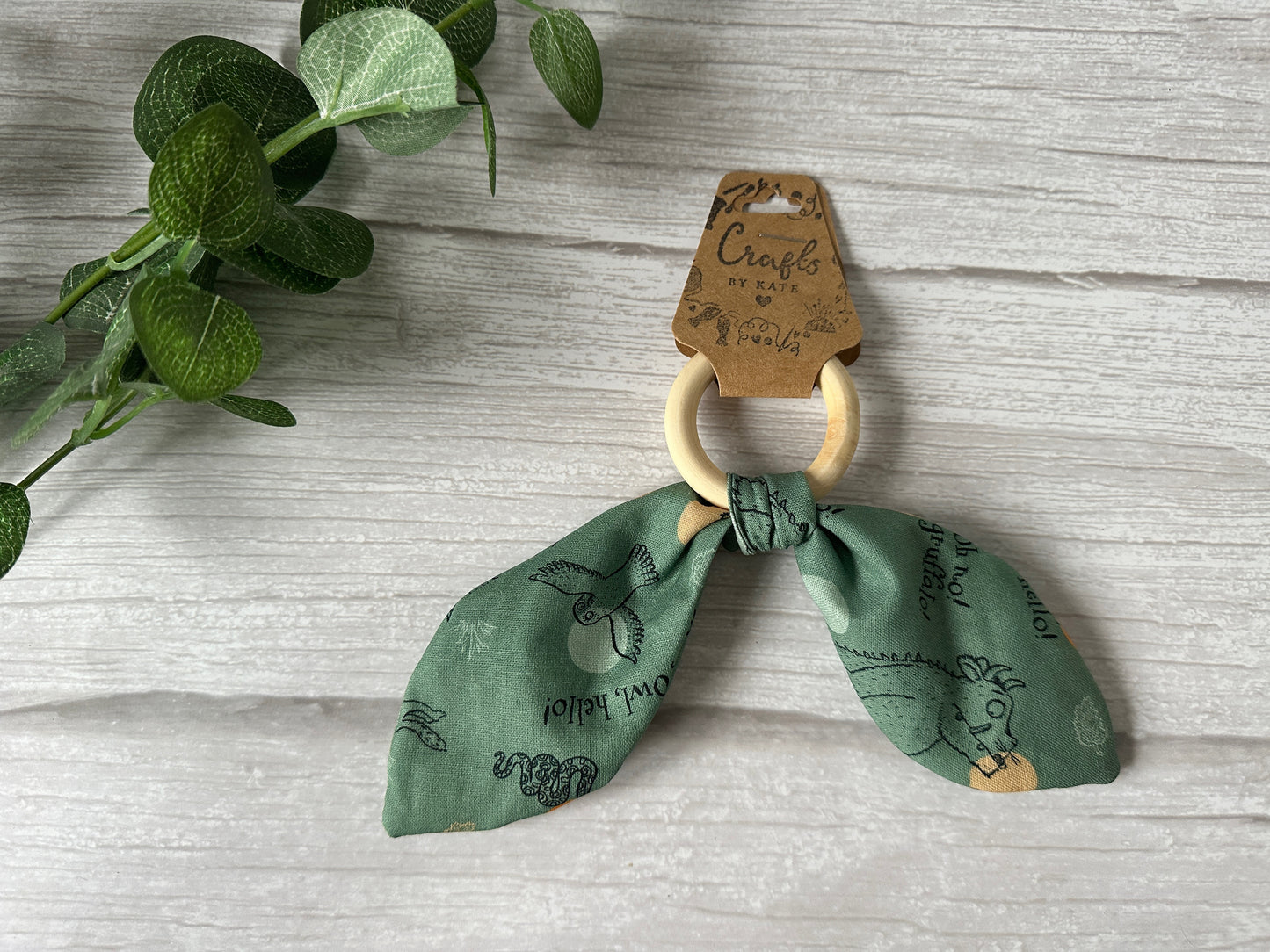 The Cotton & Bamboo Baby Teething Rattle - Green Gruffalo by Crafts by Kate features a green cotton fabric with animal patterns tied around a handcrafted teething bamboo ring, set on a light wooden surface with a craft tag and a sprig of green leaves.