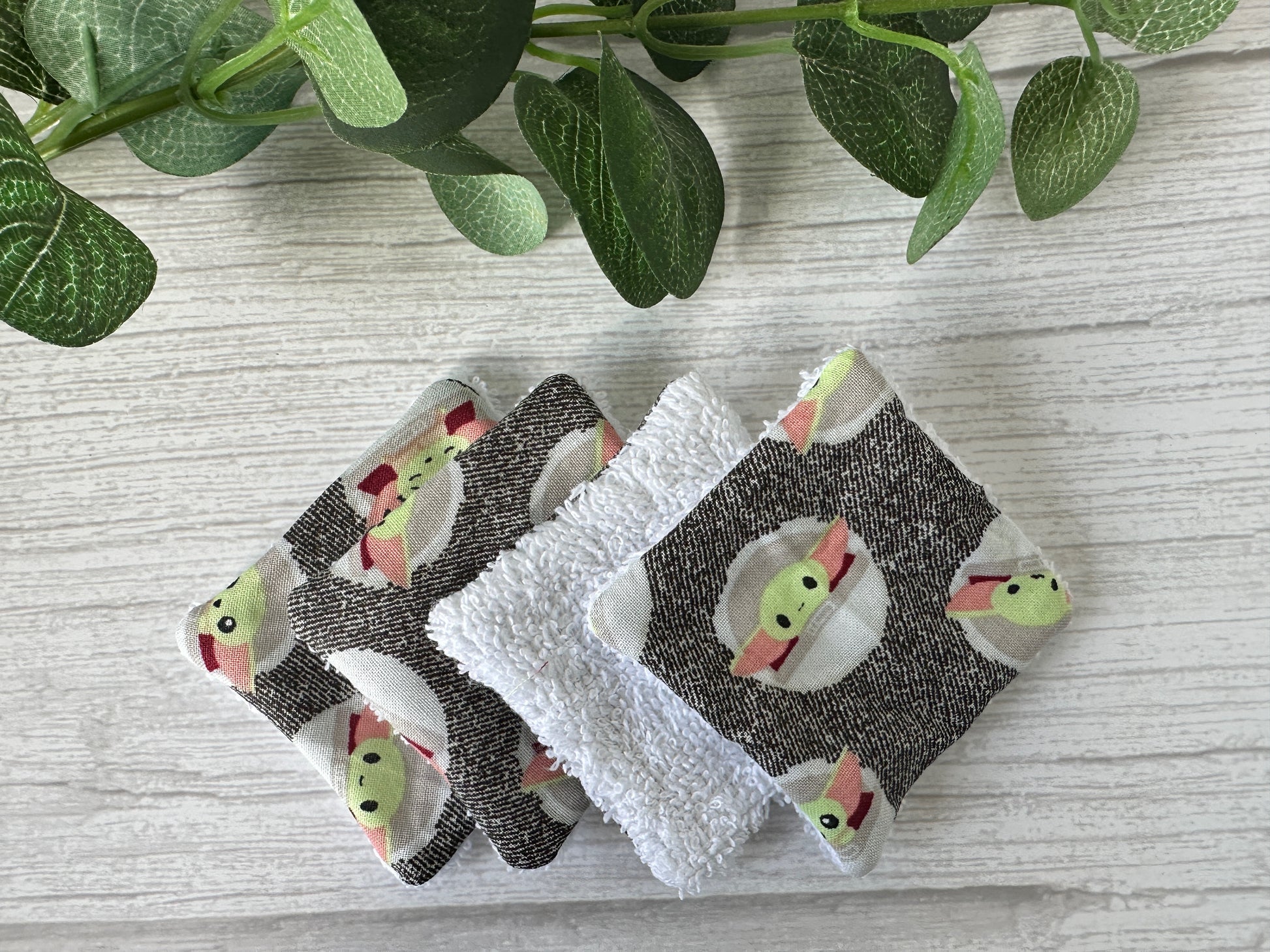 Crafts by Kate's Cotton Reusable Make-up Remover Pads - Star Wars Grogu, featuring a green alien figure, rest on a wooden surface. Made from soft white terry cloth and patterned fabric, they are eco-friendly. A sprig of green leaves can be seen in the upper left corner.