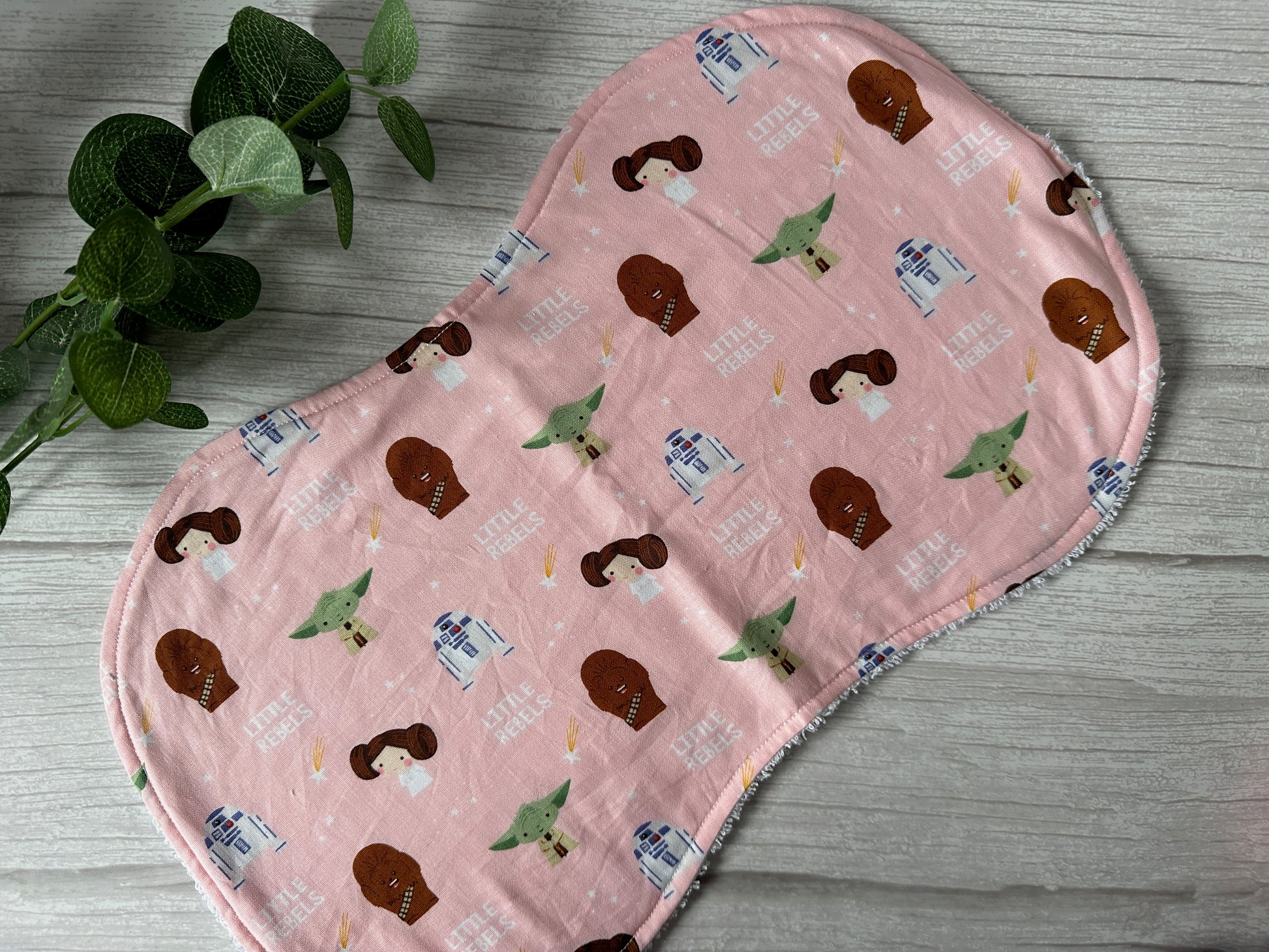 The Cotton & Terry Towelling Baby Burping Cloth, designed by Crafts by Kate, features the Little Rebels pattern with cartoon-style illustrations of Star Wars characters such as Princess Leia and Chewbacca. Made from ultra-soft cotton, it displays "LITTLE REBEL" in the background. Accompanied by a small plant, it creates a charming scene.