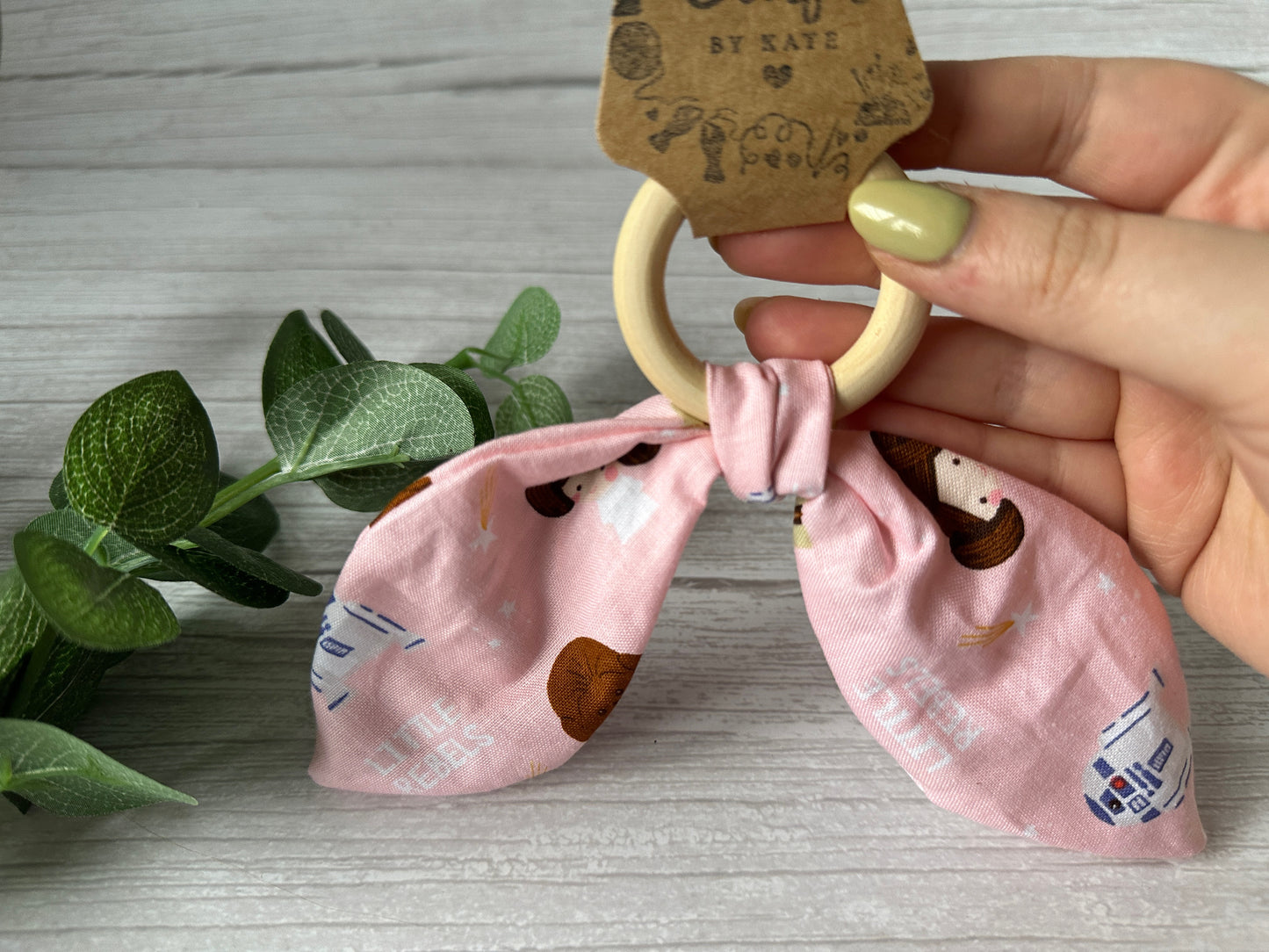 A hand with green nails holds the Crafts by Kate "Cotton & Bamboo Baby Teething Rattle - Star Wars Little Rebels," featuring a wooden bamboo ring attached to pink fabric in a bow shape. A sprig of green leaves rests on the wooden surface below.