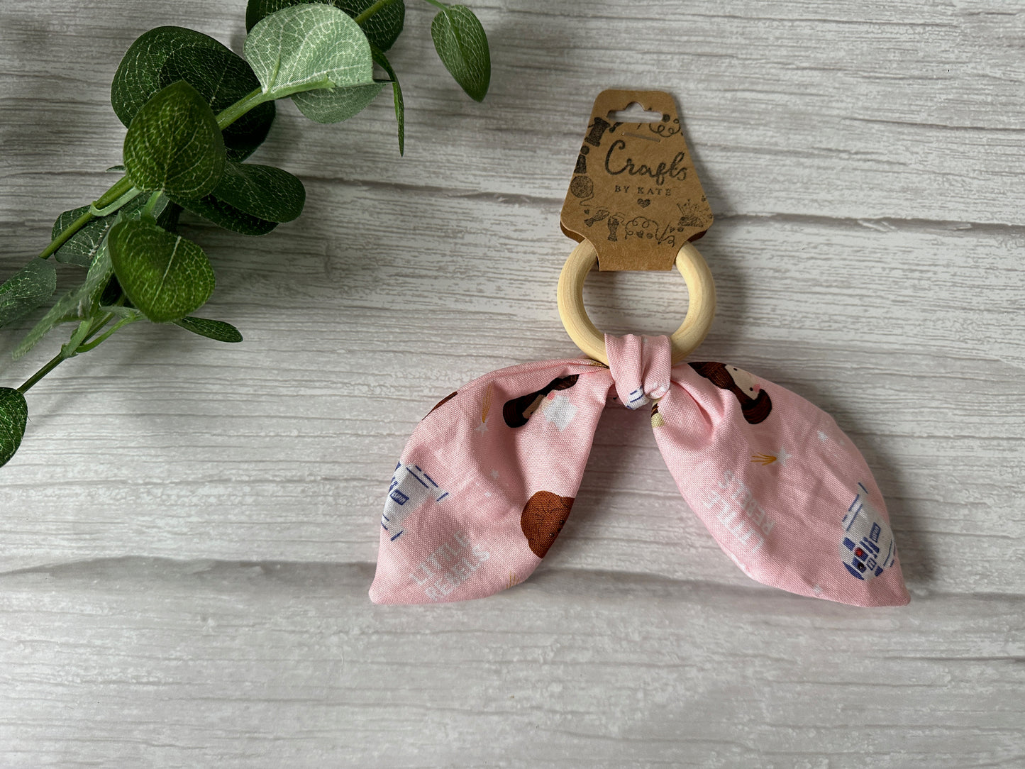 On a light wooden surface, the Crafts by Kate "Cotton & Bamboo Baby Teething Rattle - Star Wars Little Rebels" features a pink fabric bow with various patterns. Green leaves can be seen partially on the left side of the image.