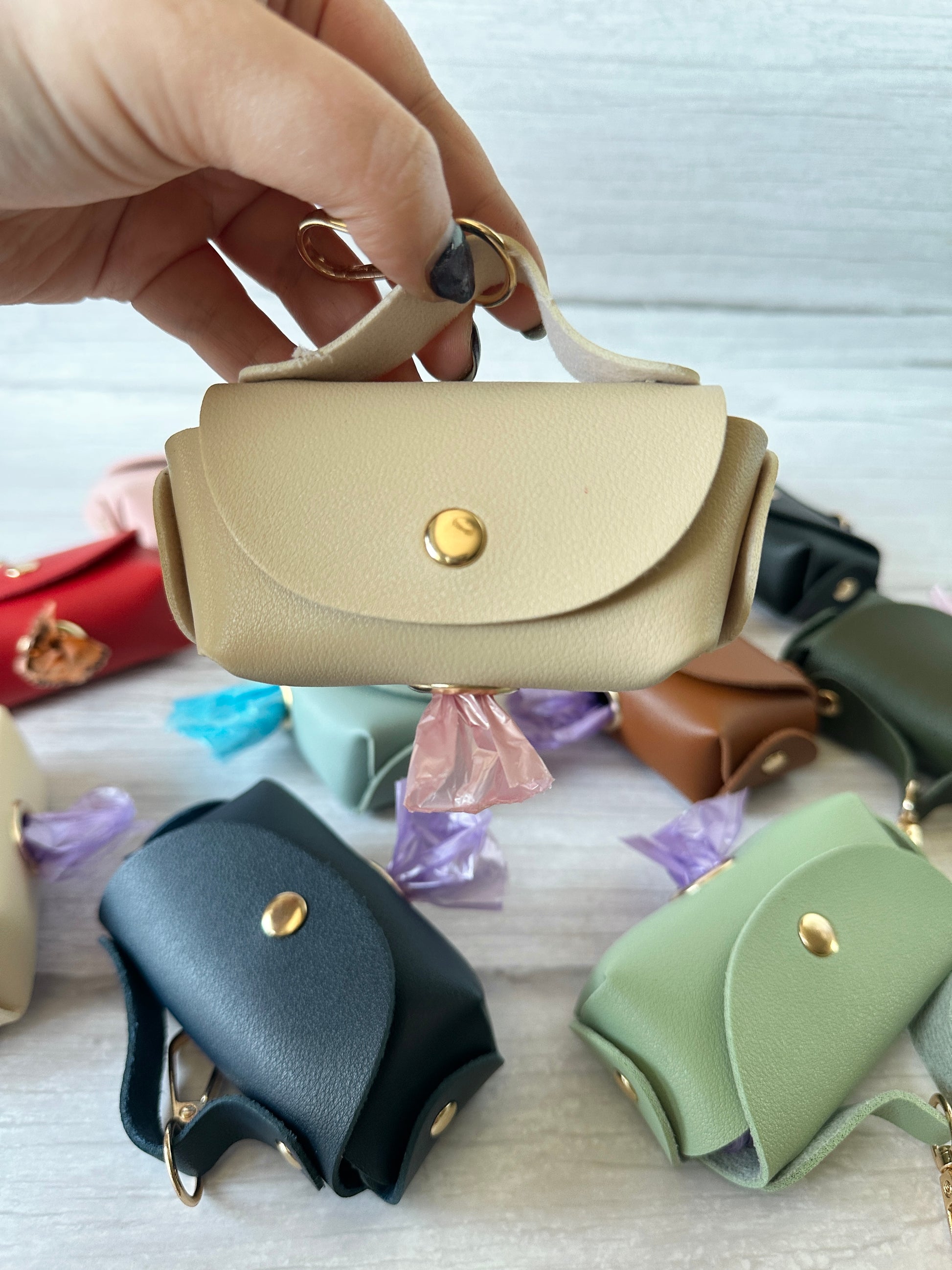 A hand holds a small beige Pet Boo Bag Holder Handbag with a gold clasp that dispenses a purple pet poo bag. Scattered across the light surface are other Crafts by Kate chic handbag-style mini bags in various colors.