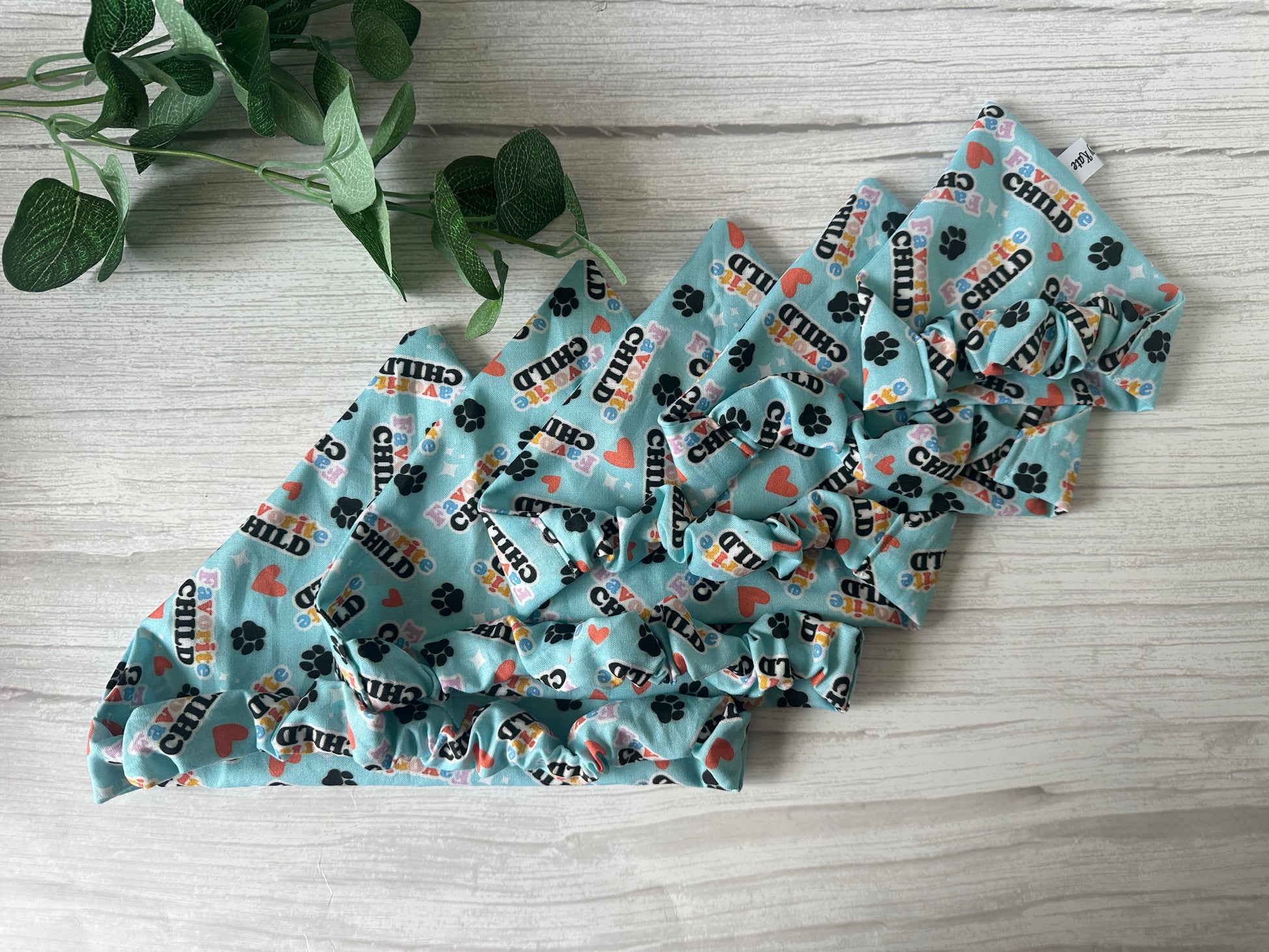 Five "Favourite Child" cotton pet scrunchie bandanas by Crafts by Kate, featuring hearts and paw prints, are neatly folded on a light wooden surface next to a small green leafy plant.