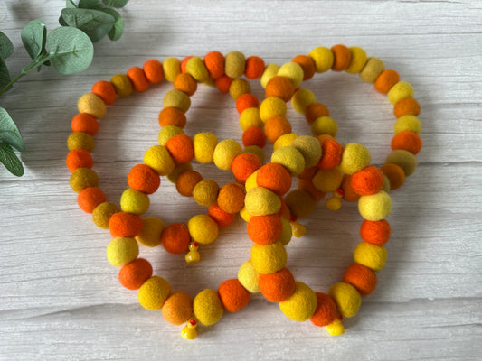 The PomPom Pet Collar - Rubber Ducks by Crafts by Kate features four beaded bracelets in orange and yellow felt, displayed on light wood—ideal as an elasticated pet collar. A sprig of green leaves graces the upper left corner.