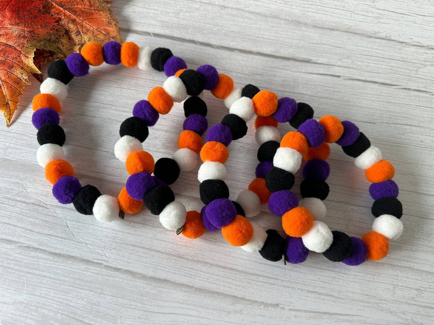 Four *PomPom Pet Collars - Halloween* by *Crafts by Kate*, featuring handcrafted, elasticated pet collars made of small, round felt balls in black, white, orange, and purple, are displayed on a wooden surface with an autumn leaf in the top-left corner. This arrangement and color scheme create a festive Halloween-themed appearance.