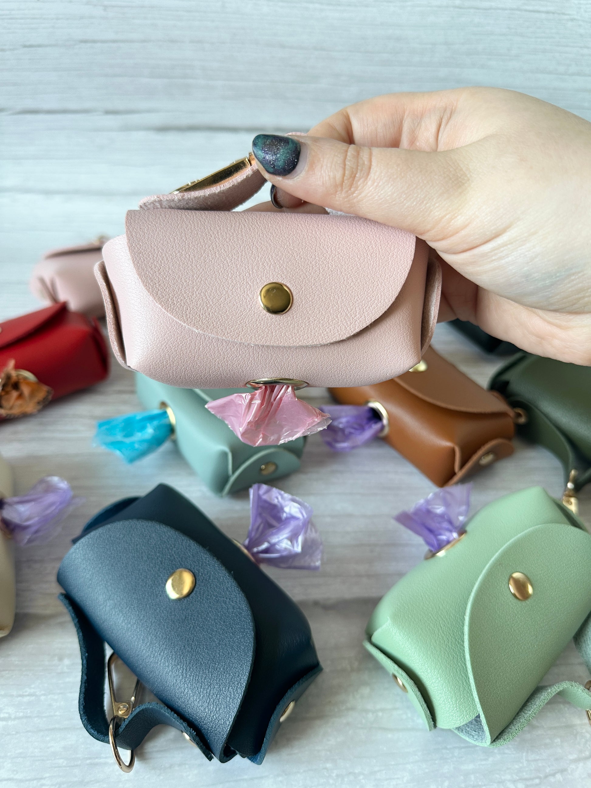 A hand holds a Crafts by Kate Pet Boo Bag Holder Handbag, a small handbag-shaped pouch in pink faux leather with a brass button. It contains lavender plastic bags. Various colors of these pouches are scattered on the wooden surface.