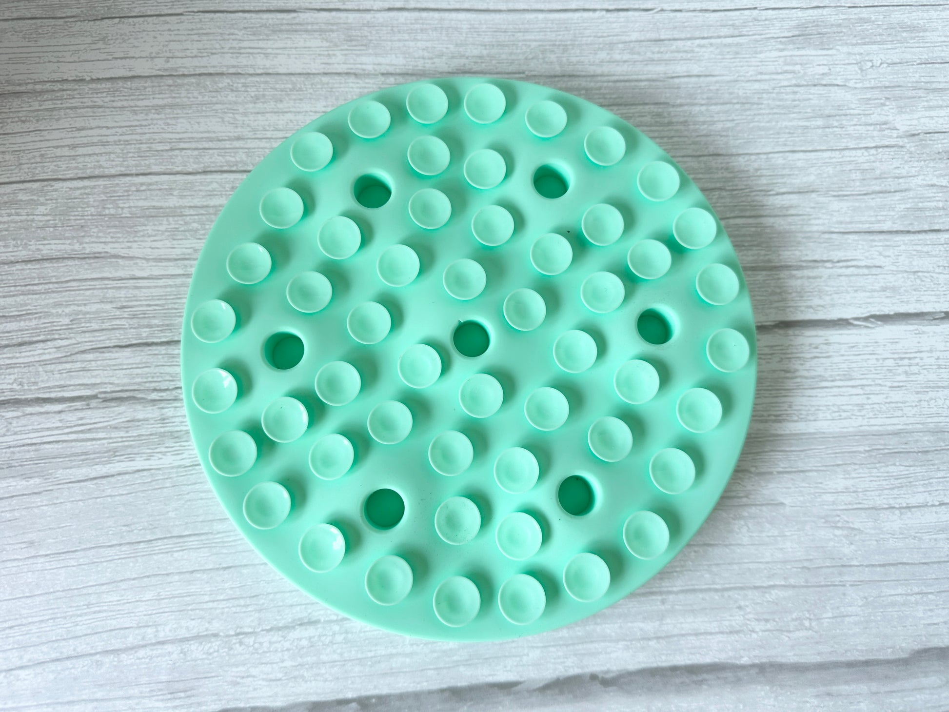 The Slow Feeder Pet Mat by Crafts by Kate is a turquoise, washable mat with evenly spaced, raised suction cups on a light wood surface; it's round with a textured pattern to aid pet digestion and keep mealtime tidy.