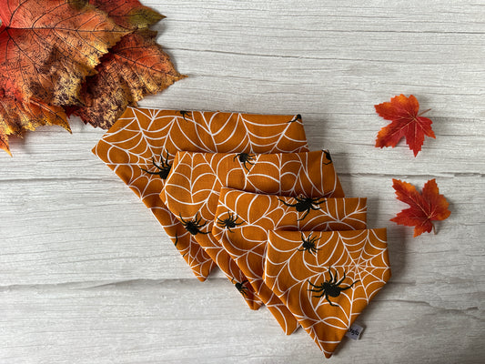 A set of (Copy) Cotton Pet Scrunchie Bandanas - Spider Vibes from Crafts by Kate are arranged fan-like on a light wooden surface, giving off serious spider web and black spider designs. Autumn leaves, in shades of red and orange, are scattered around the bandanas.