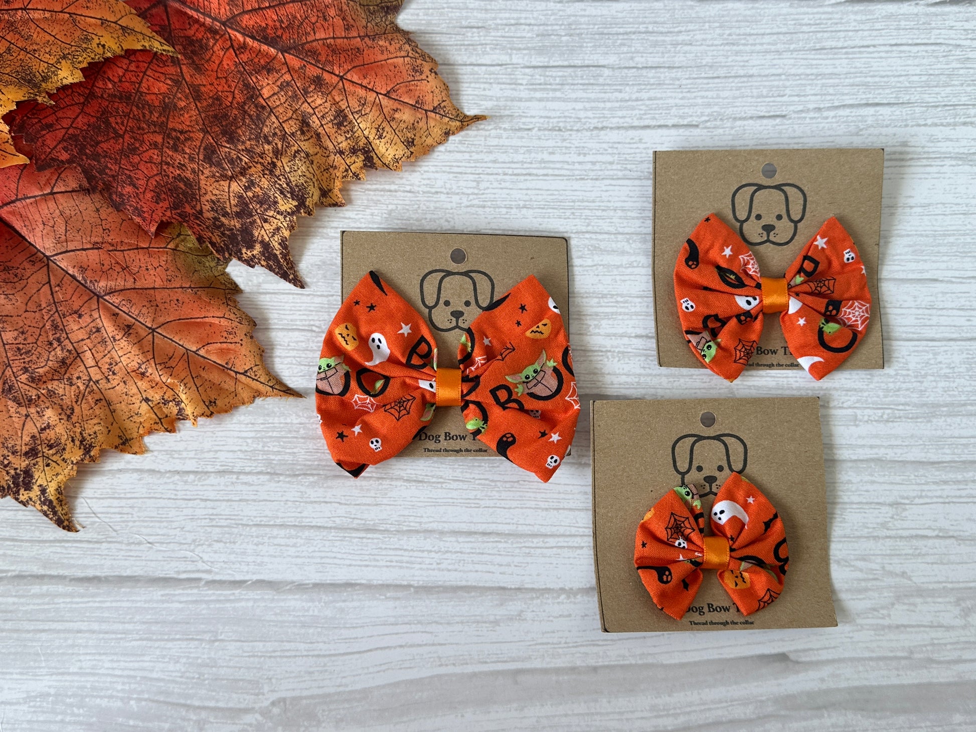 Three "Cotton Pet Bow with collar attachment - Halloween Baby Yoda" by Crafts by Kate, featuring a Halloween-themed design with hints of Baby Yoda on orange fabric, are displayed on brown packaging with a dog illustration. These bows are set on a light wooden surface next to autumn leaves.