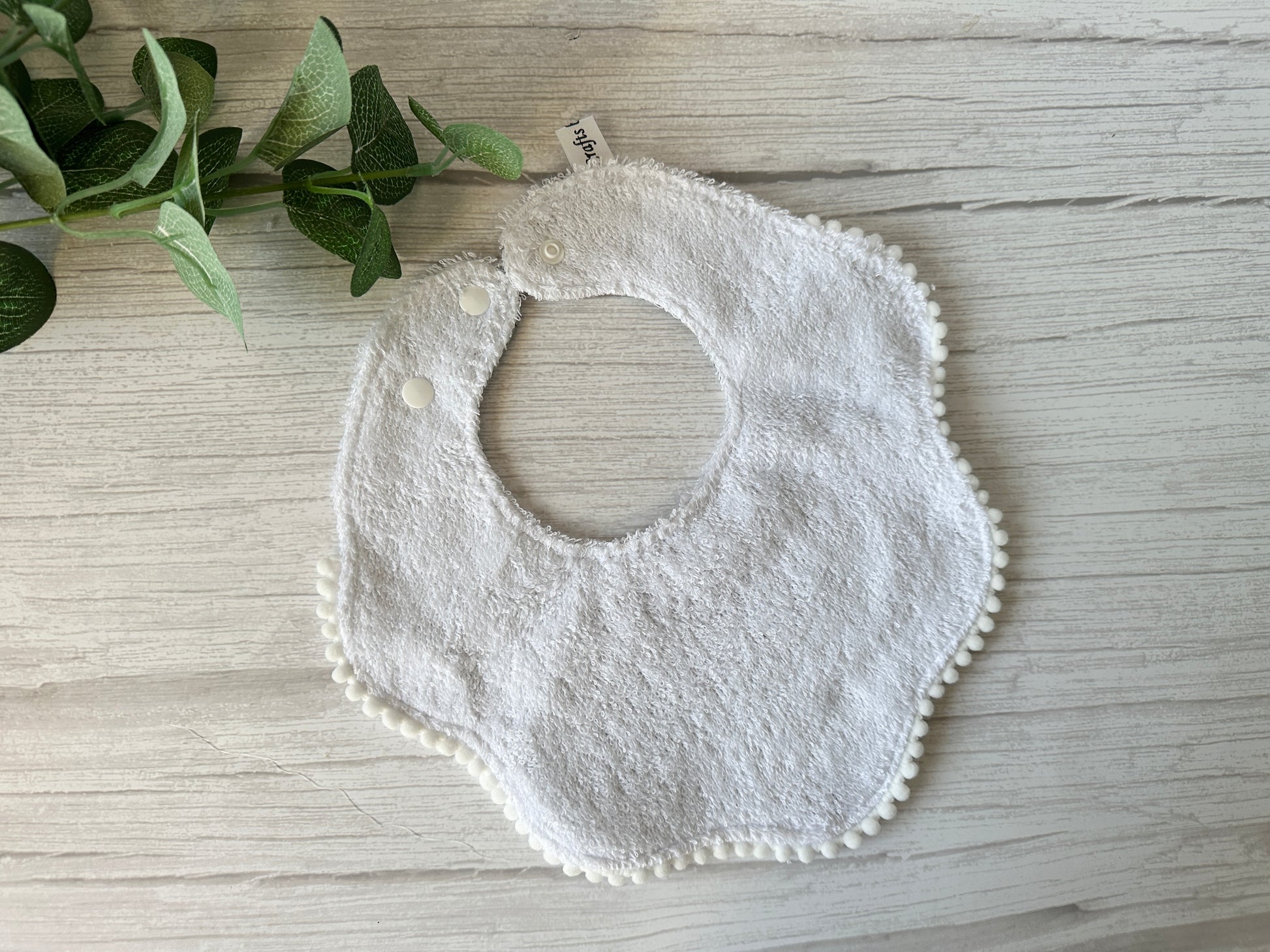 Crafts by Kate's Cotton & Terry Towelling Baby Gift Set - Floral Bambi, featuring a white textured bib with scalloped edges and pom-pom trim, lies on a light wooden surface. A green leafy plant in the top left corner completes this perfect baby gift set addition.