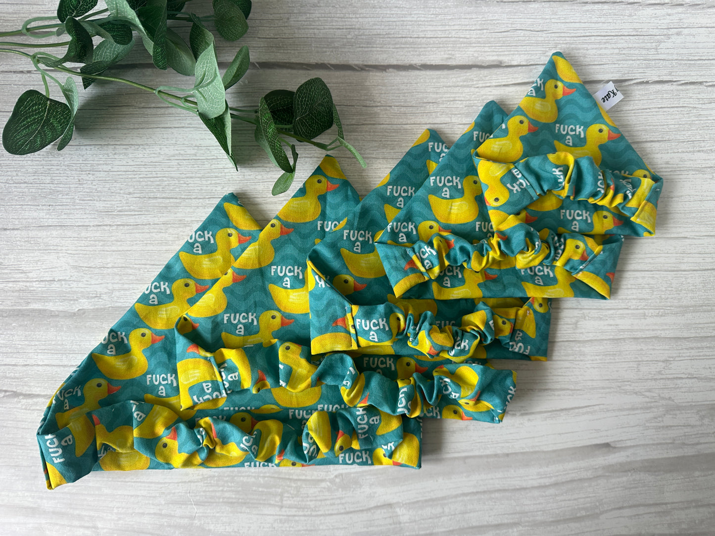 Four Crafts by Kate Cotton Pet Scrunchie Bandanas and four matching bows with yellow rubber duck prints and "F*** a duck" are displayed on a light wood surface next to a green leafy plant.