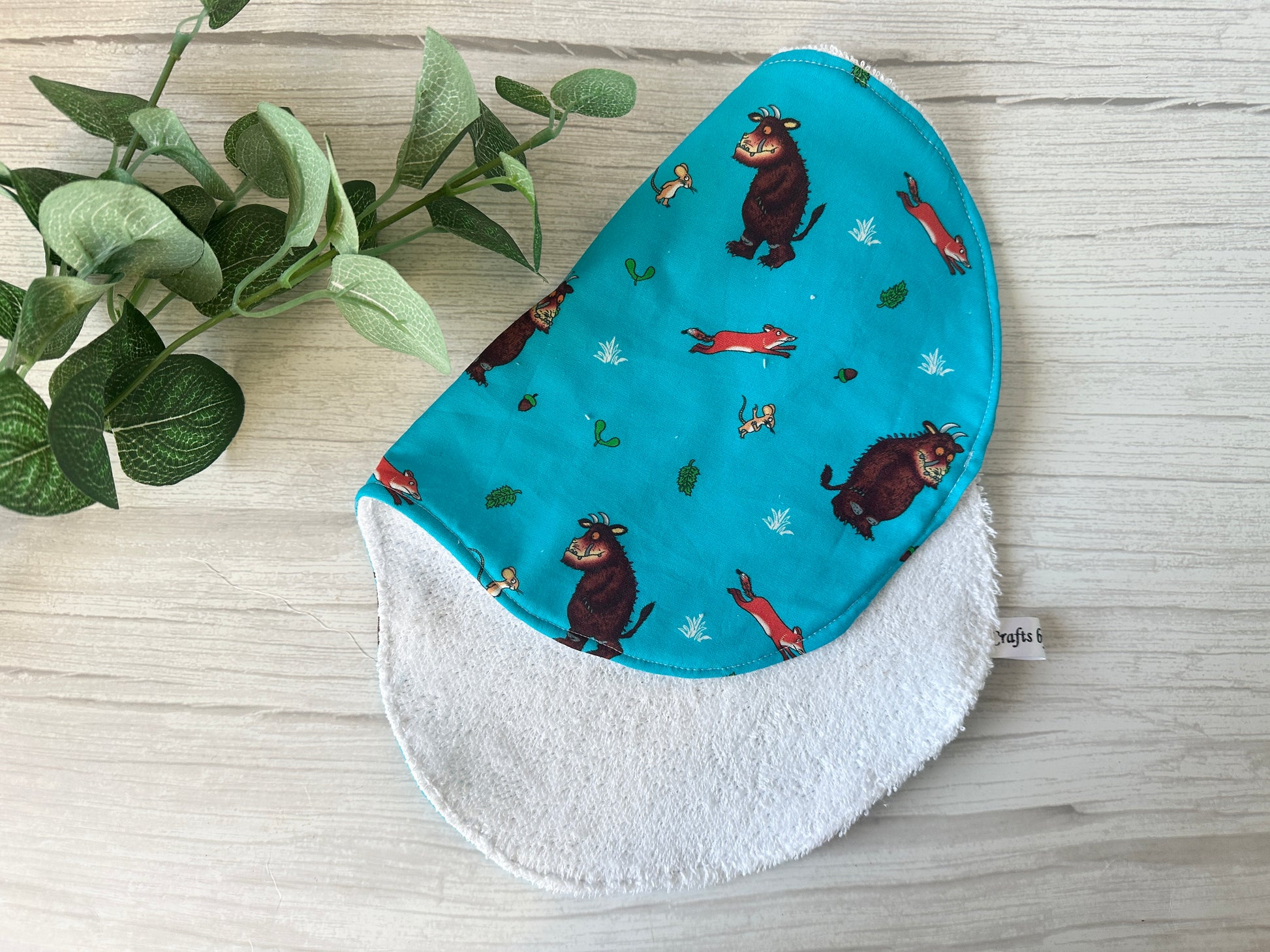 The "Cotton & Terry Towelling Baby Gift Set" by Crafts by Kate features an ultra-soft bib with turquoise fabric adorned with cartoon animals such as a brown bear and red fox, with a textured white reverse. Nearby, a green potted plant enhances the gentle ambiance on the light wood surface.