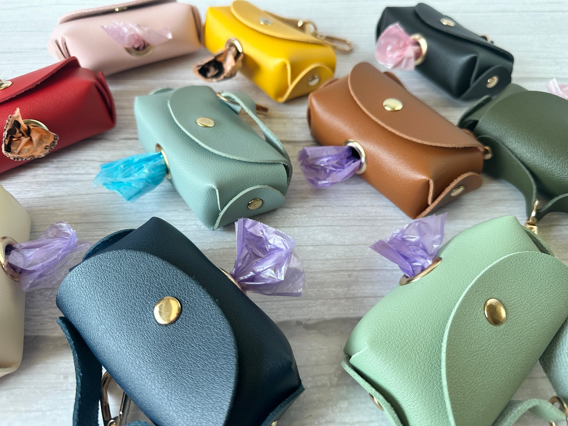 Handbag-shaped faux leather pouches by Crafts by Kate, in a circle on light wood, offer vibrant colors: red, pink, yellow, green, blue, and brown. Each Pet Poo Bag Holder Handbag features a button closure and dispenses a colored waste bag for easy pet clean-up.