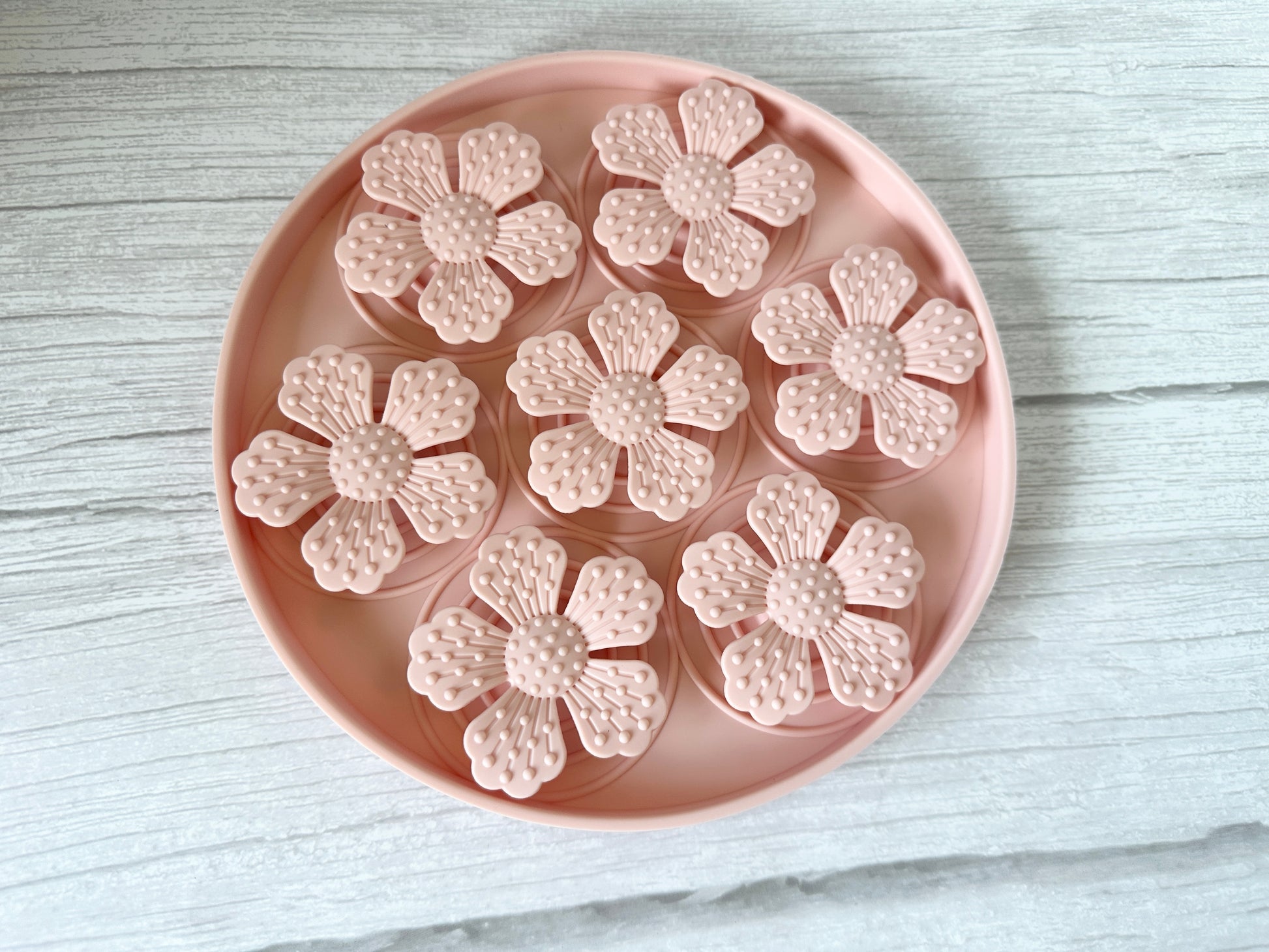 Seven floral-shaped objects sit on a round tray, their textured light pink petals are delicately arranged, atop the Crafts by Kate Slow Feeder Pet Mat, a white wood-like surface that enhances the design's simplicity and elegance.