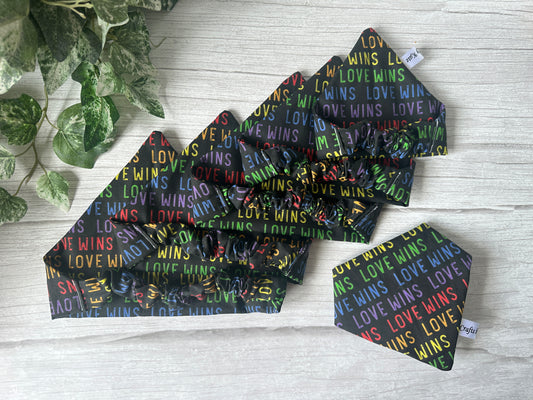 The Cotton Pet Scrunchie Bandana - Love Wins by Crafts by Kate features rainbow "LOVE WINS" on black cotton, celebrating the LGBTIQA+ community. Handcrafted accessories are styled on a light wooden surface with green leaves in the corner.