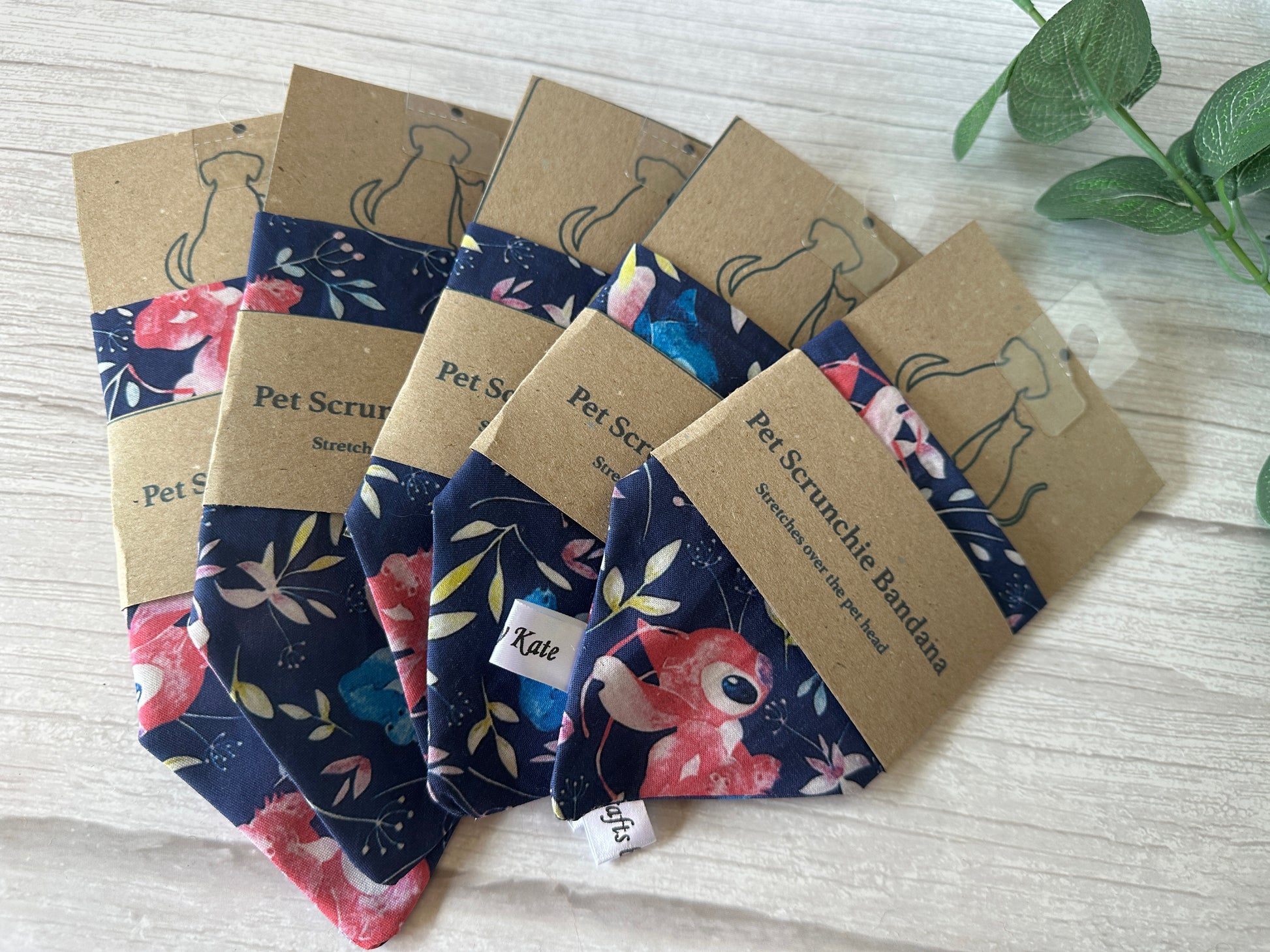 Crafts by Kate's Stitch Obsession collection features handcrafted cotton pet scrunchie bandanas with floral patterns on a wooden surface. Each is wrapped in a kraft paper holder with a dog illustration, and a sprig of green leaves decorates the top right corner.