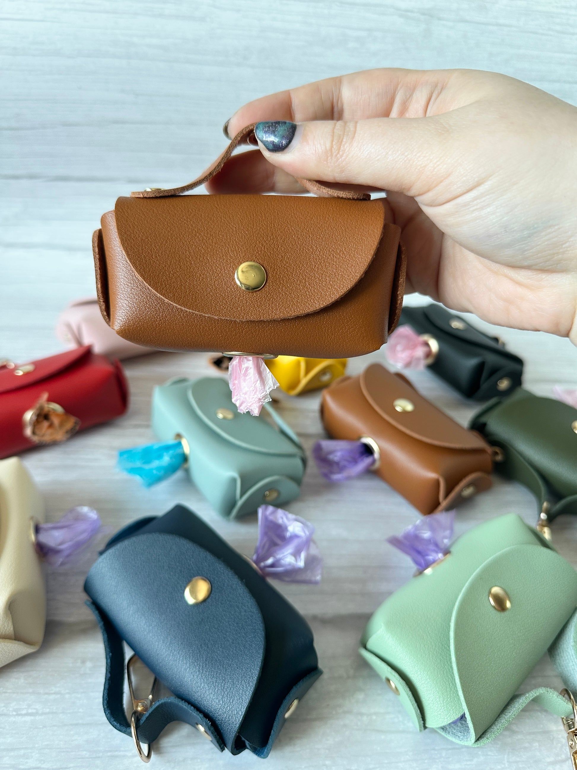 A hand holds a Pet Boo Bag Holder Handbag by Crafts by Kate, a small brown faux leather pouch with a snap closure and loop handle. It dispenses a purple pet poo bag. Similar pouches in various colors are scattered on a light surface in the background.