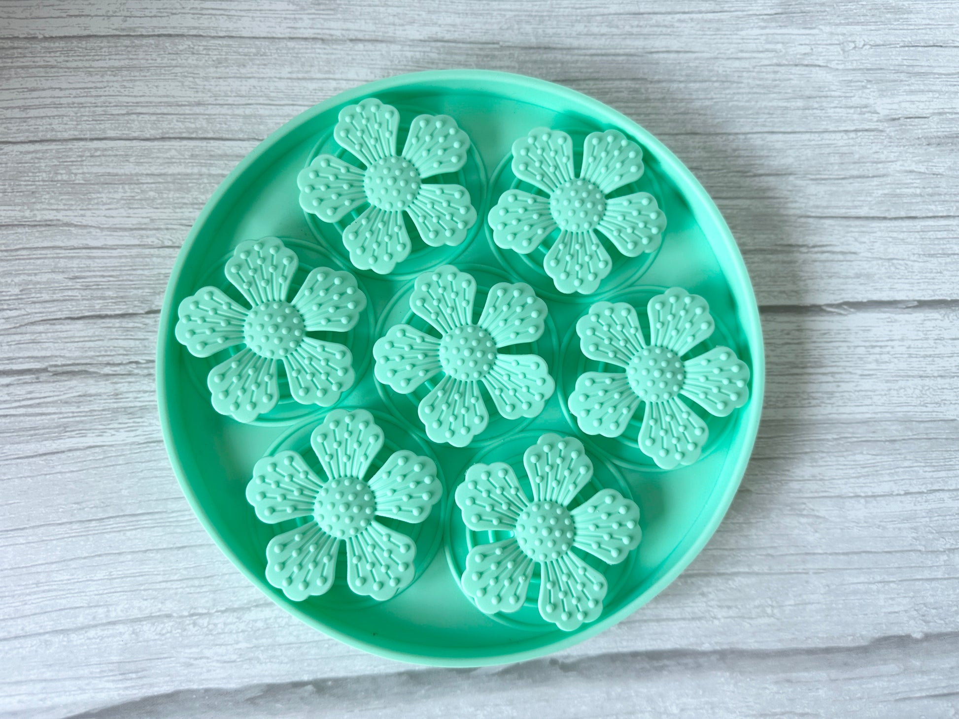 The Crafts by Kate Slow Feeder Pet Mat is a turquoise circular tray with suction cups featuring six flower-shaped molds, each with detailed petal and center designs. It securely rests on a textured white wooden surface and is washable.